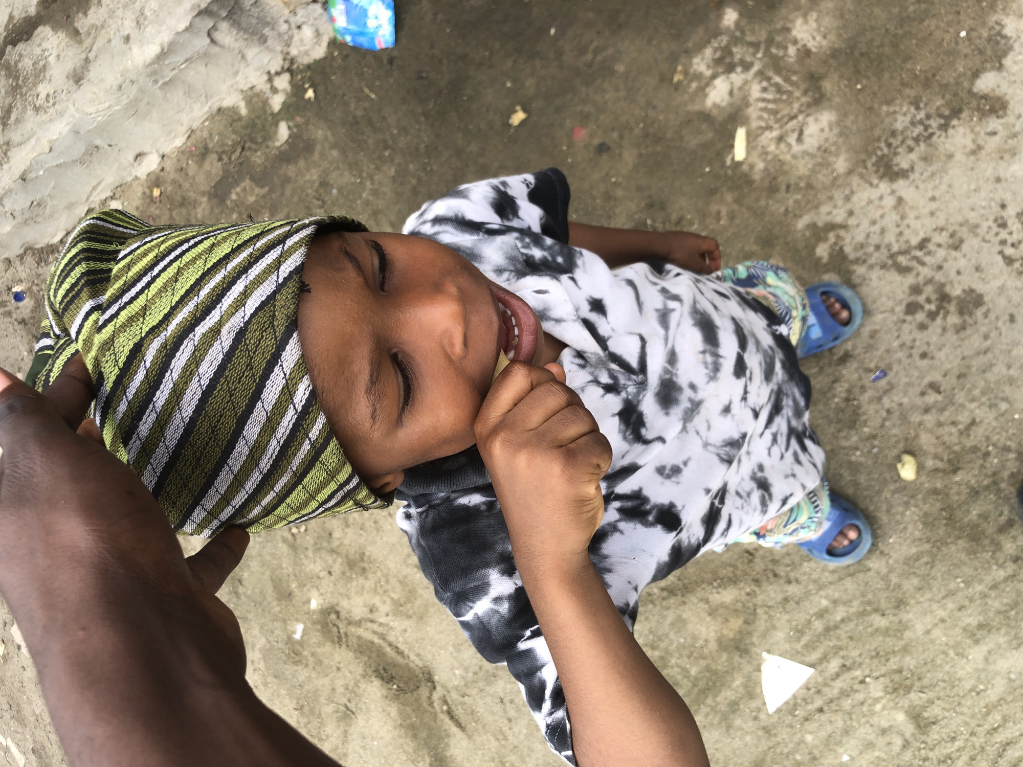 An overhead shot shows a young child in the city of Ado, Nigeria, wearing a black and white tie-dye shirt and a green, white, and black striped hat. The child is eating something and has their hand to their mouth. They are also wearing blue slides. An adult's hand is visible at the top of the frame, appearing to adjust the child's hat. The ground is composed of dirt and concrete.
