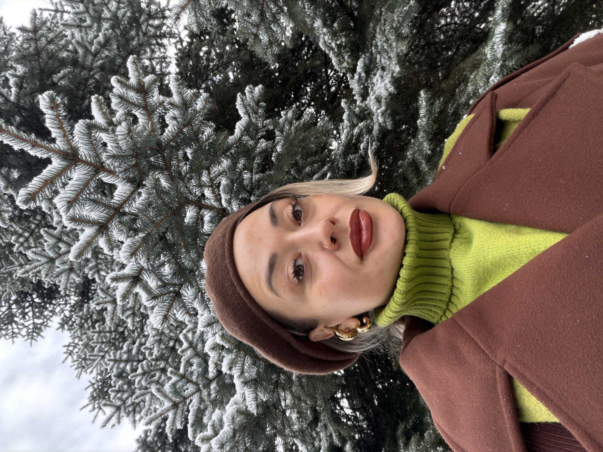 A woman is centered in the frame, wearing a brown beret, a lime green turtleneck sweater, and a brown coat. She has fair skin, light blonde hair peeking out from under the beret, and is wearing gold hoop earrings. Her lips are coated in a deep burgundy lipstick, and she has a nose ring. She appears to be taking a selfie.
The background consists of a dense evergreen tree covered in snow. The image is taken outdoors during daylight hours. Based on the presence of evergreen trees covered in snow, this could be a park or natural area within Krasnoyarsk, Russia. Specific landmarks within the city are not discernible.