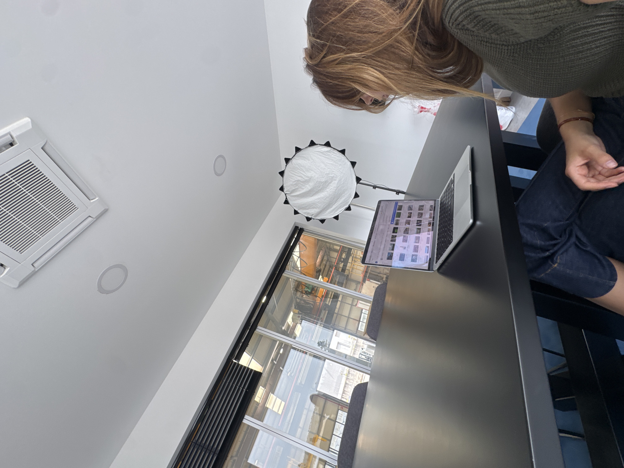Captured from a low-angle perspective, the image depicts an indoor scene, likely an office, studio, or co-working space, with a person leaning over a dark-colored table. The person, with long brown and blonde hair, is wearing a green knitted sweater and blue denim jeans, and appears to be focused on a silver MacBook Pro laptop. The laptop screen displays a grid of various image thumbnails.
To the left, a white air conditioning unit is mounted on the white ceiling, alongside two recessed circular lights. A large, circular white photography reflector with a black frame stands on a pole in the background.
A prominent feature is the large window spanning the lower-middle section of the image, offering a view of an industrial or commercial area outside. Through the window, parts of large machinery, possibly cranes or industrial structures, and other buildings are visible under bright daylight, suggesting the photo was taken during the day. The reflections on the window also show parts of the interior space. The overall ambiance is modern and functional. No specific discernible text is visible other than the faint image thumbnails on the laptop screen.