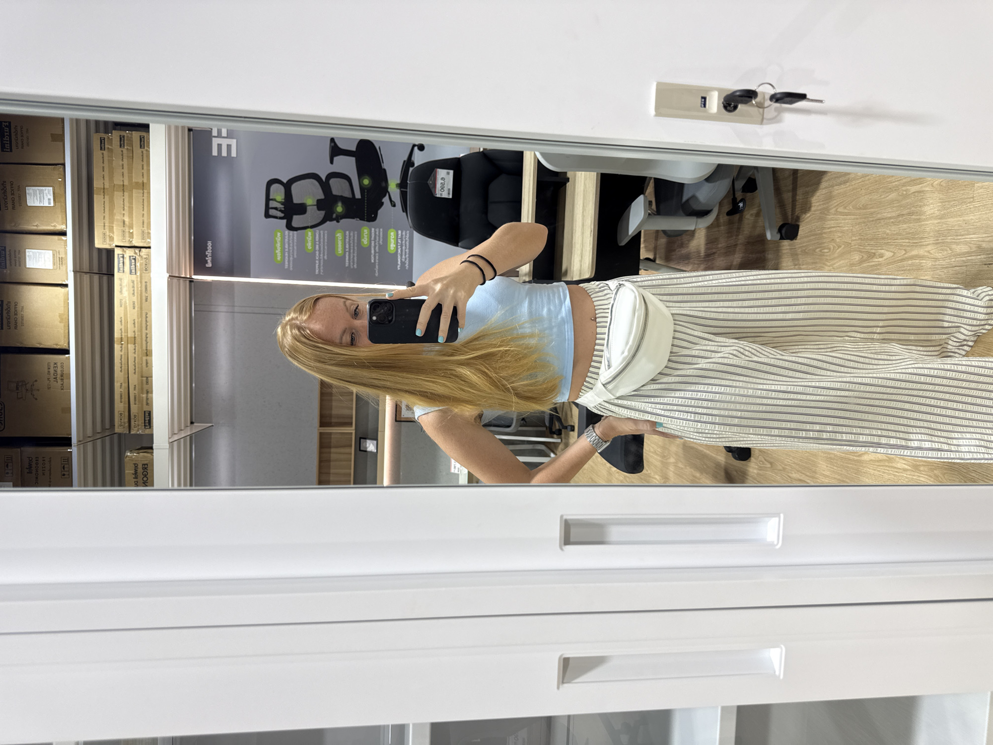 A young woman with blonde hair takes a mirror selfie in a retail store. She is wearing a light blue crop top, striped wide-leg pants, and a white fanny pack. Her left hand is on her hip, and she is holding a black smartphone in her right hand. Behind her, the store displays office chairs and shelving units with boxes. The flooring is light wood. The mirror is large and framed by a white structure, possibly a wardrobe or cabinet. The location is indicated as Baan Chaweng Noi, Thailand.