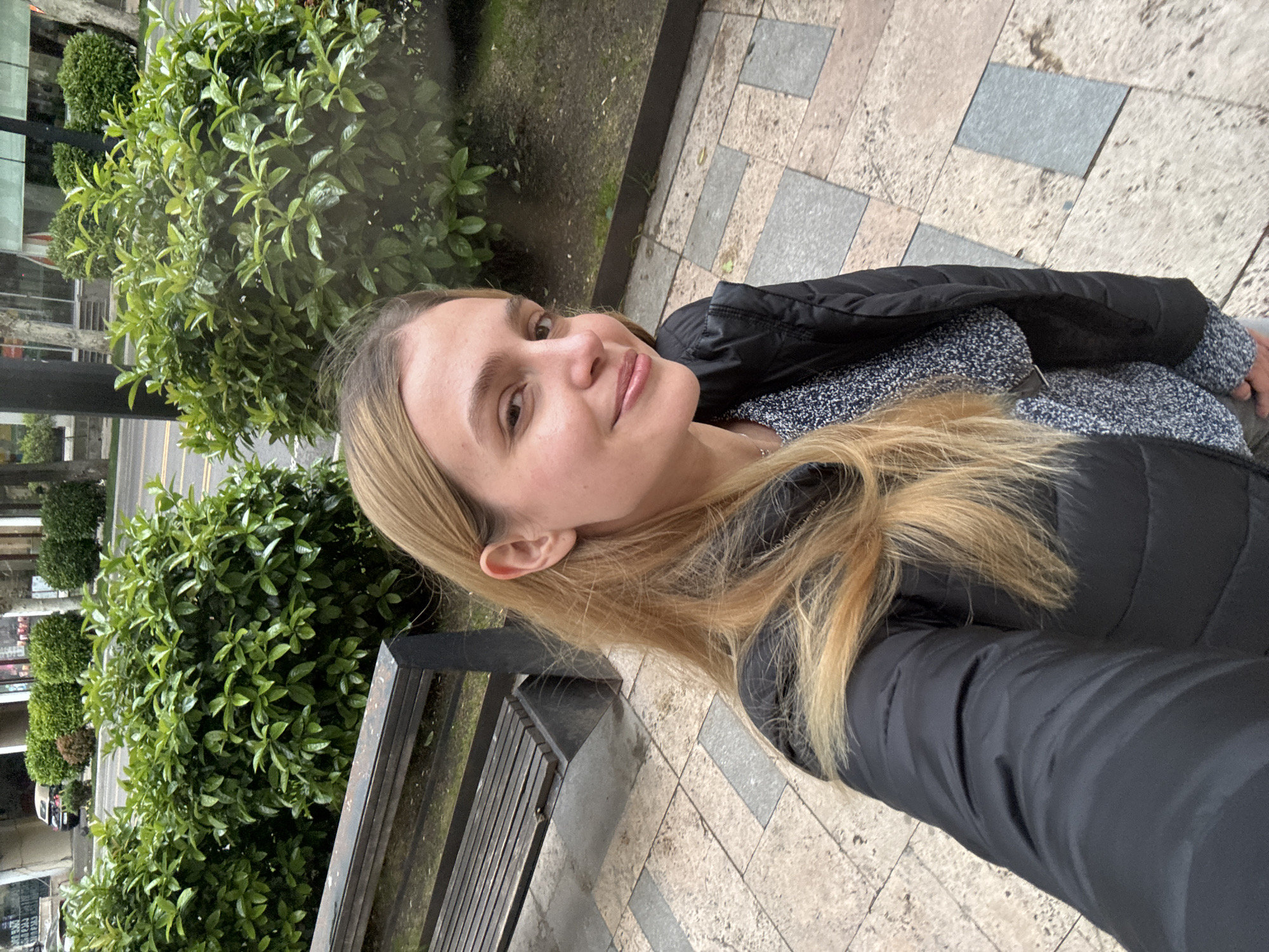 A selfie photo shows a fair-skinned woman with light blonde hair, a black puffer jacket, and a grey sweater. She is outside on a sidewalk made of light-colored square tiles. Behind her are green bushes, a bench with metal slats, and buildings across the street, suggesting a street in a city. The location is most likely in Tbilisi, Georgia, based on the provided context.