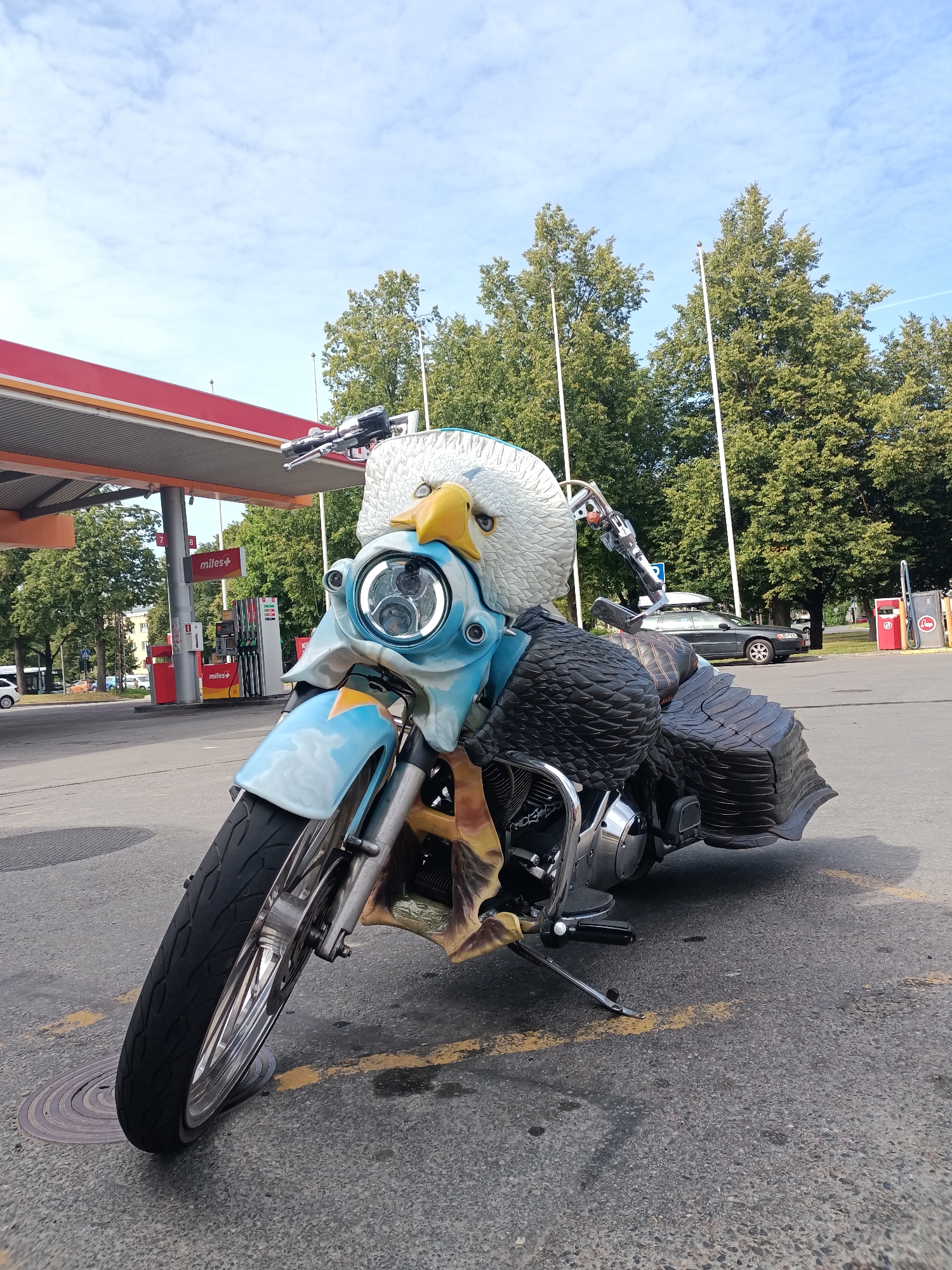 The image depicts a custom-built motorcycle with an eagle-themed design, parked at a Miles+ gas station in Tallinn, Estonia. The motorcycle features an elaborate front fairing shaped like an eagle's head, with a yellow beak and blue and white feather details. The saddlebags are designed to resemble large eagle feathers. The motorcycle is parked on asphalt, with yellow lines on the ground. In the background, there is a red and white gas station canopy with several gas pumps. Trees and buildings are visible behind the gas station, and a dark-colored Volvo station wagon with a roof box is parked in the background. The sky is partly cloudy with blue patches. There are no people visible in the image.