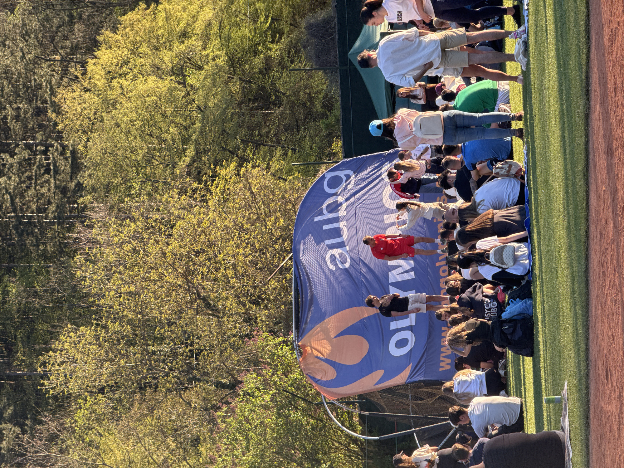 A large group of people is gathered on a grassy area, possibly a sports field, in Blagoevgrad, Bulgaria. In the background, trees line the edge of the field. A large banner with the text "OLYMPIC" is positioned in front of the group. Many people are sitting, kneeling, or standing. The image is rotated counter-clockwise.
