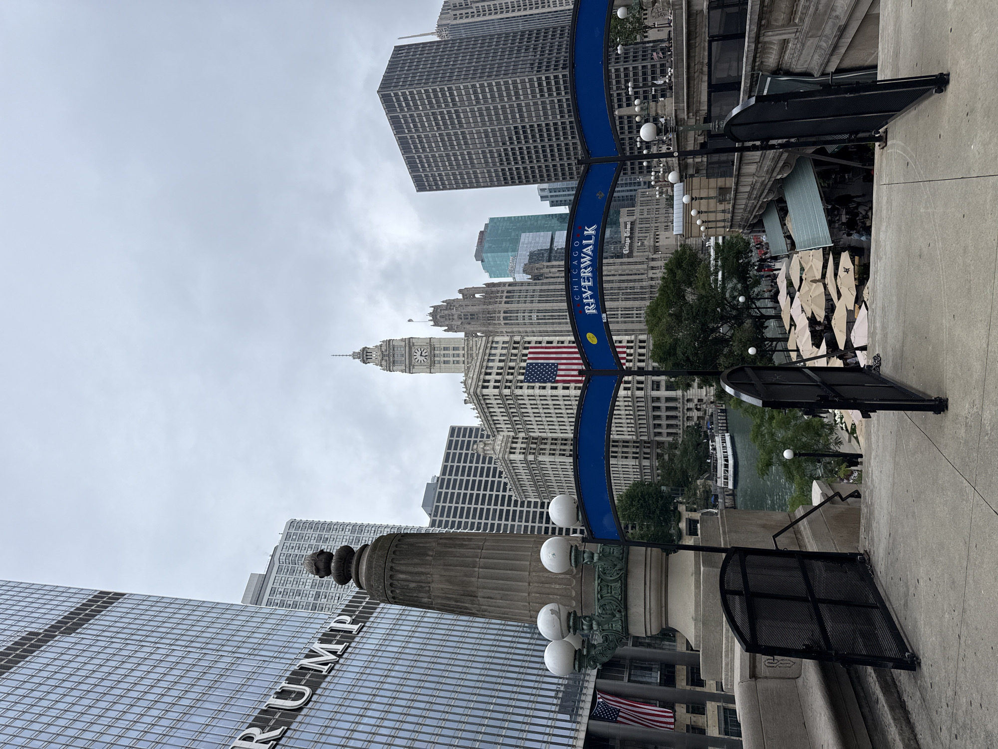 The image captures a view of the Chicago Riverwalk on an overcast day. The scene features a street level perspective looking towards several skyscrapers and a bridge. The Chicago River is visible in the background, with a tour boat navigating its waters. The Riverwalk area appears to be set up for outdoor dining, with many umbrellas folded and stacked, suggesting a break in service or preparation for changing weather.
Prominently displayed is a blue arched sign that reads "CHICAGO RIVERWALK." An American flag is visible on the side of a building in the midground. The Trump International Hotel & Tower is identifiable by the large, reflective glass facade with the partial text "TRUMP" visible. Other buildings in the background vary in architectural style, from modern glass structures to older, more ornate buildings. A clock tower is also visible on one of the older buildings.
The weather appears cloudy and slightly hazy, contributing to a muted color palette. The overall mood is somewhat subdued due to the overcast conditions. The perspective suggests the viewer is on a walkway or a set of stairs leading down towards the Riverwalk. There are black metal fences and lampposts lining the walkway. The scene is a typical urban landscape showcasing the city's architecture and its popular waterfront attraction.