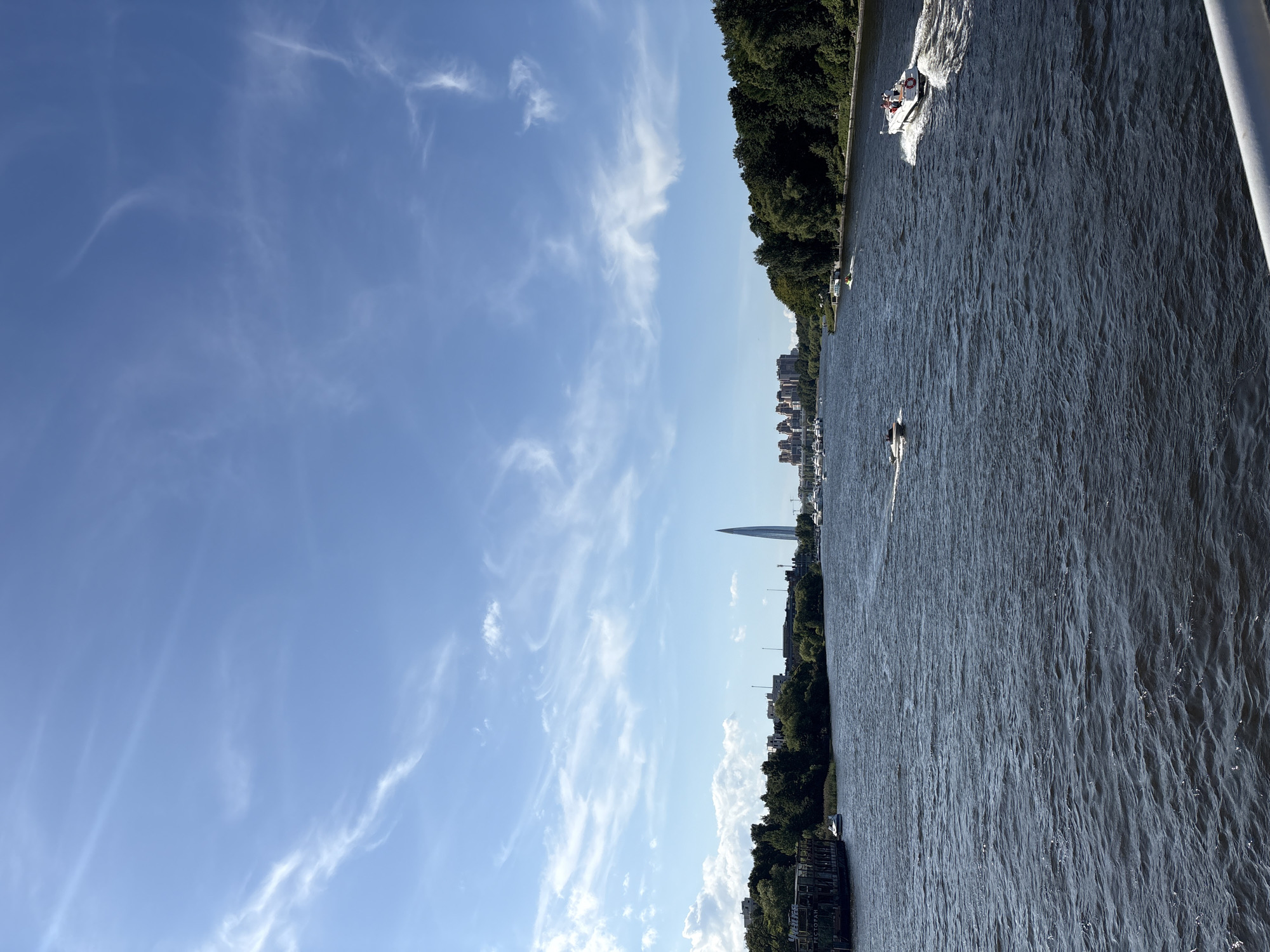 A wide, dark blue river with a rippling surface dominates the foreground of the image. Two white speedboats are visible on the water. One is closer to the right side of the frame, moving away from the viewer and leaving a white wake. It carries at least three people. Another speedboat is further left and closer to the center, moving towards the left side of the frame.
The riverbanks are lined with lush green trees. On the left bank, a large, dark, boat-like structure with signs is docked. In the distance, across the river, a modern, slender skyscraper with a distinctive pointed top is visible against a bright blue sky filled with wispy white clouds. Further to the left of the skyscraper, a cluster of reddish-brown buildings with multiple stories can be seen. The overall scene depicts a sunny day in an urban or semi-urban environment with a prominent waterway and modern architecture in the background.