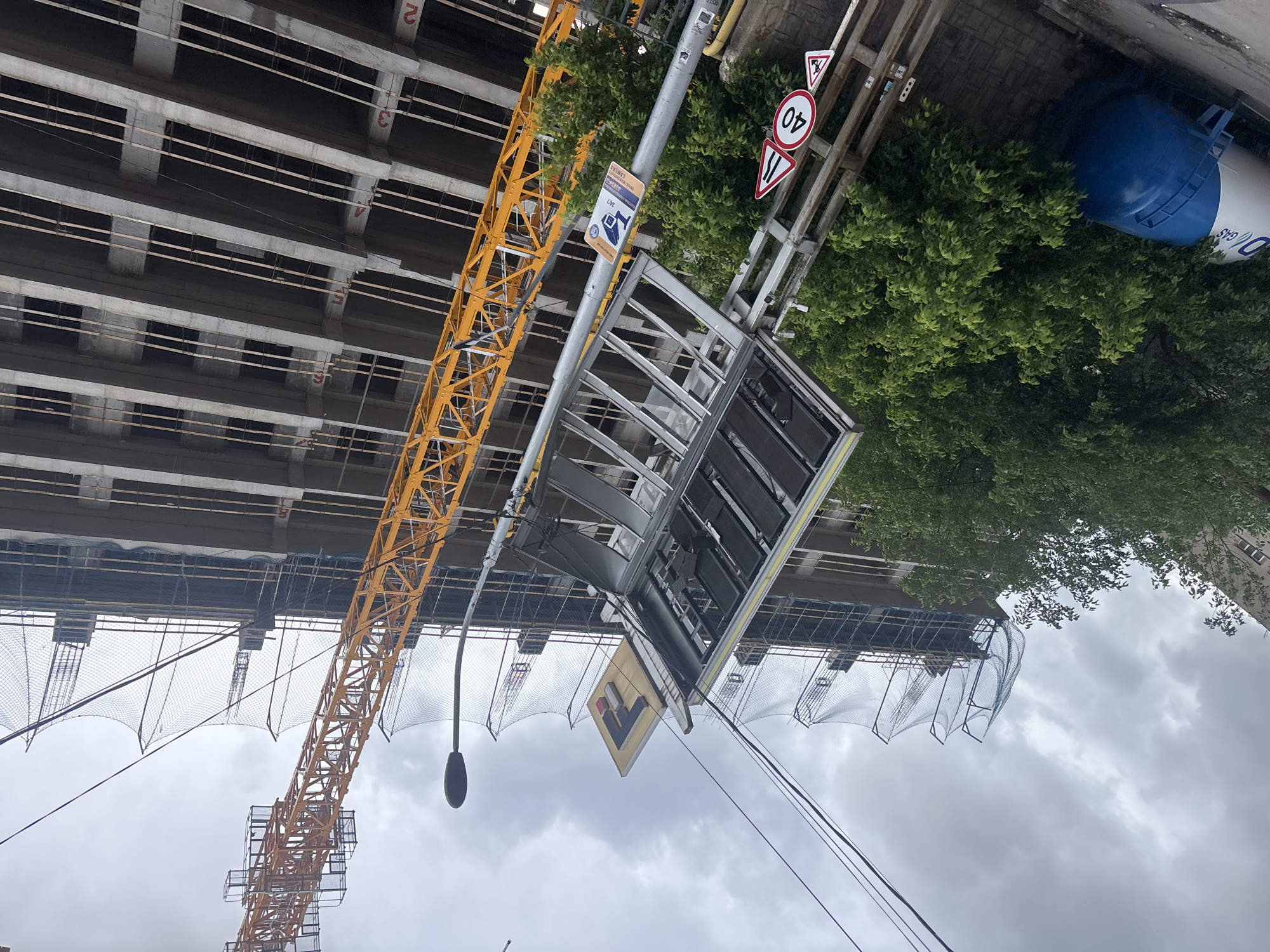 The image depicts a construction site in Tbilisi, Georgia. A tall, yellow crane stands adjacent to a multi-story concrete building under construction. The building's floors are visible, marked with numbers "2" and "3". A pedestrian overpass with signage, including a speed limit sign (40 km/h), crosses a street. A blue, cylindrical tank is visible among the trees. The sky is overcast. A street lamp is visible.
