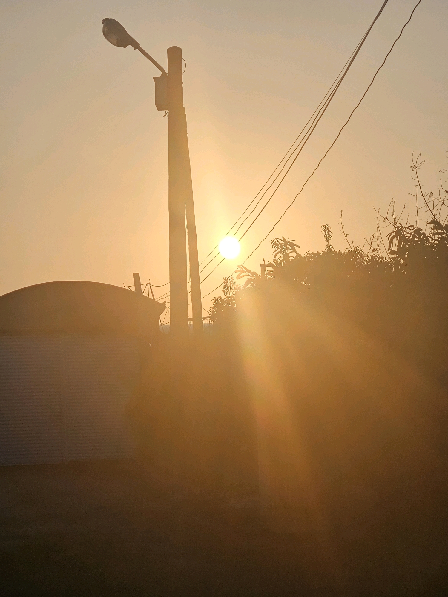 The image depicts a sunset scene in Huwei Township, Taiwan. The sun, a bright orb, is visible in the upper right quadrant of the frame, casting strong golden rays and lens flare across the image. In the foreground, a ribbed, dome-shaped structure, possibly a silo or storage tank, is partially visible on the left. A utility pole with a streetlamp and electrical wires stretches vertically through the center of the image, silhouetted against the bright sky. Trees and vegetation are visible behind the utility pole and to the right, their forms blurred by the intense light and atmospheric haze. The overall impression is one of a rural or semi-industrial setting bathed in the warm glow of a setting sun.