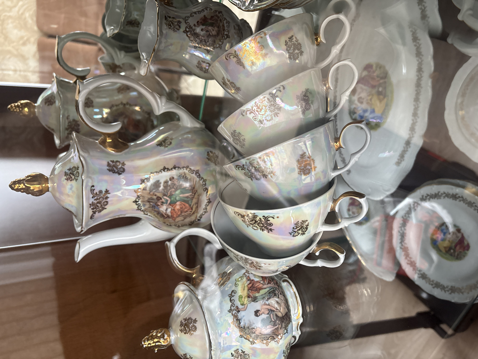 An ornate, vintage tea set, likely made of porcelain or ceramic, is the central subject of this indoor photograph. The set features a lustrous, iridescent pearlescent glaze, giving it a shimmering quality. It is intricately decorated with extensive gold gilding in floral and scroll motifs, reminiscent of damask patterns. Several pieces also showcase colorful, Rococo-style vignettes depicting classical figures in pastoral or mythological scenes, rendered in muted tones of green, orange, and blue.
Visible components of the tea set include a teapot with a gilded finial and handle, numerous teacups with elegant, often gilded, handles, and matching saucers. The pieces are arranged on a highly reflective, glossy surface, possibly glass, creating prominent mirror images beneath and around them. The teacups are stacked, suggesting they are either on display in a cabinet or storage, or arranged for viewing in a shop.
The background is softly blurred, revealing vague shapes and lighter tones, possibly indicating a room interior. There is no specific activity taking place, as the items are static display pieces. No text is visible within the image. The time of day or external weather conditions cannot be determined from this indoor setting. While the image itself does not contain visual cues for Krasnoyarsk, Russia, the context provided indicates this is the location where the media file was captured. The overall impression is one of elegance and antique craftsmanship.