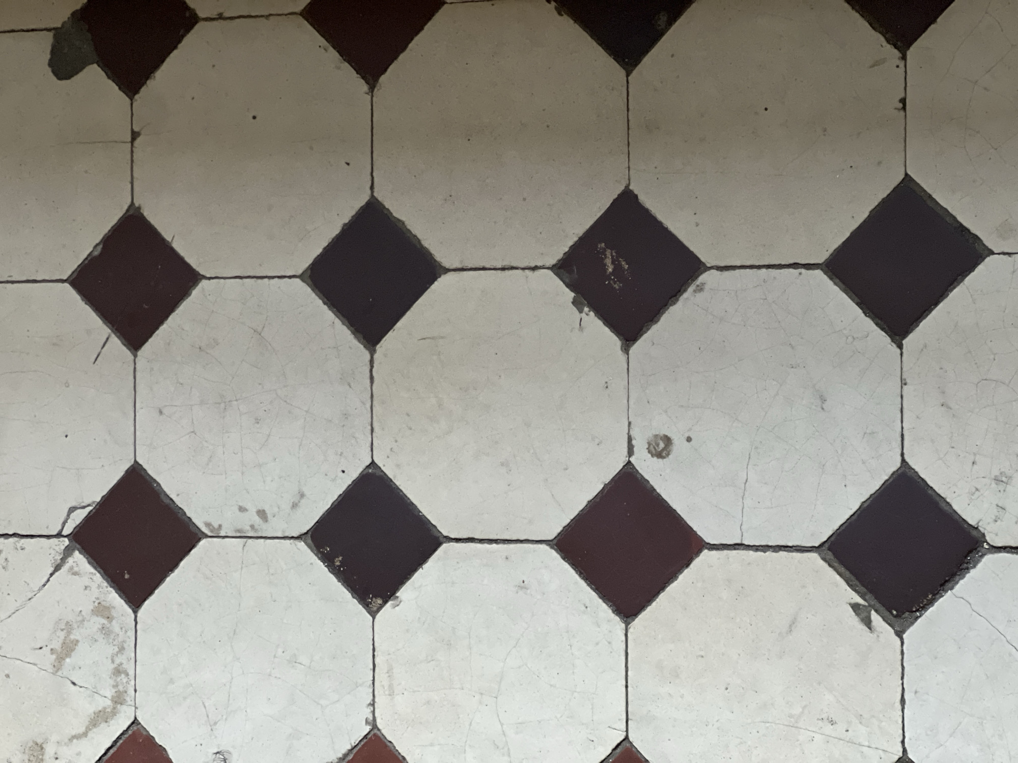 Close-up view of an old tile floor. The floor consists of alternating octagonal white tiles and square dark brown tiles. The tiles are arranged in a grid pattern. The white tiles are cracked and stained, indicating wear and age. The grout lines between the tiles are thin and dark. The floor appears to be in a state of disrepair. This type of tile pattern is commonly found in older buildings in Saint Petersburg, Russia.