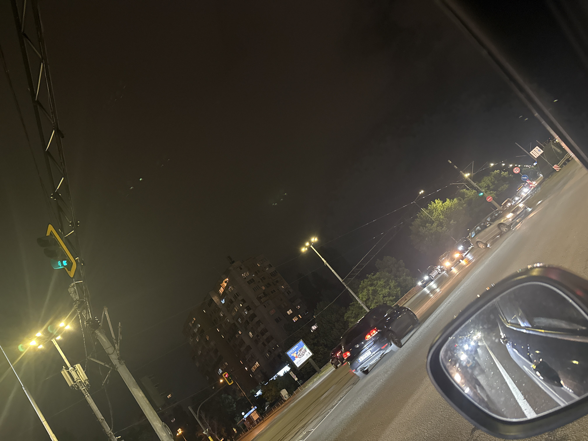 The image presents a nocturnal view of a city street in Sofia, Bulgaria, captured from inside a vehicle, likely waiting at a traffic light. The scene is illuminated by numerous artificial light sources, including streetlights, car headlights, and taillights.
In the immediate foreground, the right-side mirror of the vehicle is prominently featured, reflecting the lights of cars behind. Ahead, multiple cars are queued on a multi-lane road, their red taillights creating streaks of light on the asphalt. To the left, a green traffic light with a left-turn arrow is visible, mounted on an overhead structure, alongside bright streetlights.
In the mid-ground, a tall, multi-story apartment building with numerous lit windows stands out, characteristic of urban architecture. Trees with green foliage line the street, partially obscuring elements further in the background. A large billboard with an advertisement is also visible, though its text is illegible.
Notable details include a circular road sign displaying "70," indicating a speed limit. The license plate of a car directly in front is partially visible, showing characters consistent with Bulgarian plates, such as "E 7xxxxT." The dark night sky covers the upper portion of the image. The overall impression is that of a busy urban environment after dark, with the hustle and bustle of traffic.