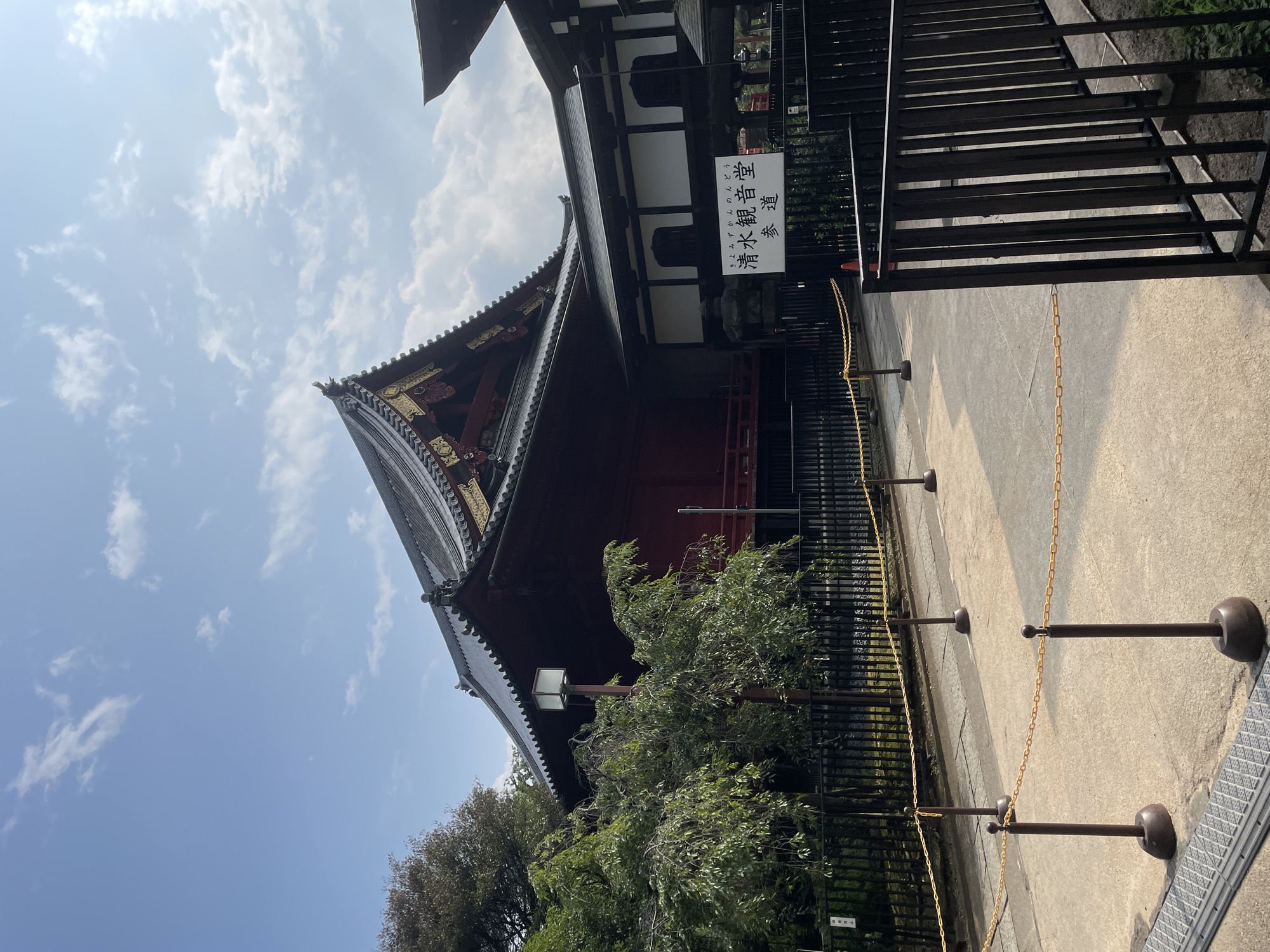 The image shows a side view of a traditional Japanese temple in Taito, Japan. The temple features a dark-tiled roof with ornate gold embellishments and a large, dark red wooden structure with an elevated platform. A white sign with Japanese text is visible in front of the temple, indicating it is the "Kiyomizu Kannon-do" and the pathway is the "Sando" (approach). The temple is surrounded by a black metal fence and lush green trees. A concrete pathway leads towards the temple, marked by stanchions and yellow chains to guide visitors. The sky above is blue with scattered white clouds. The lighting suggests it is daytime.