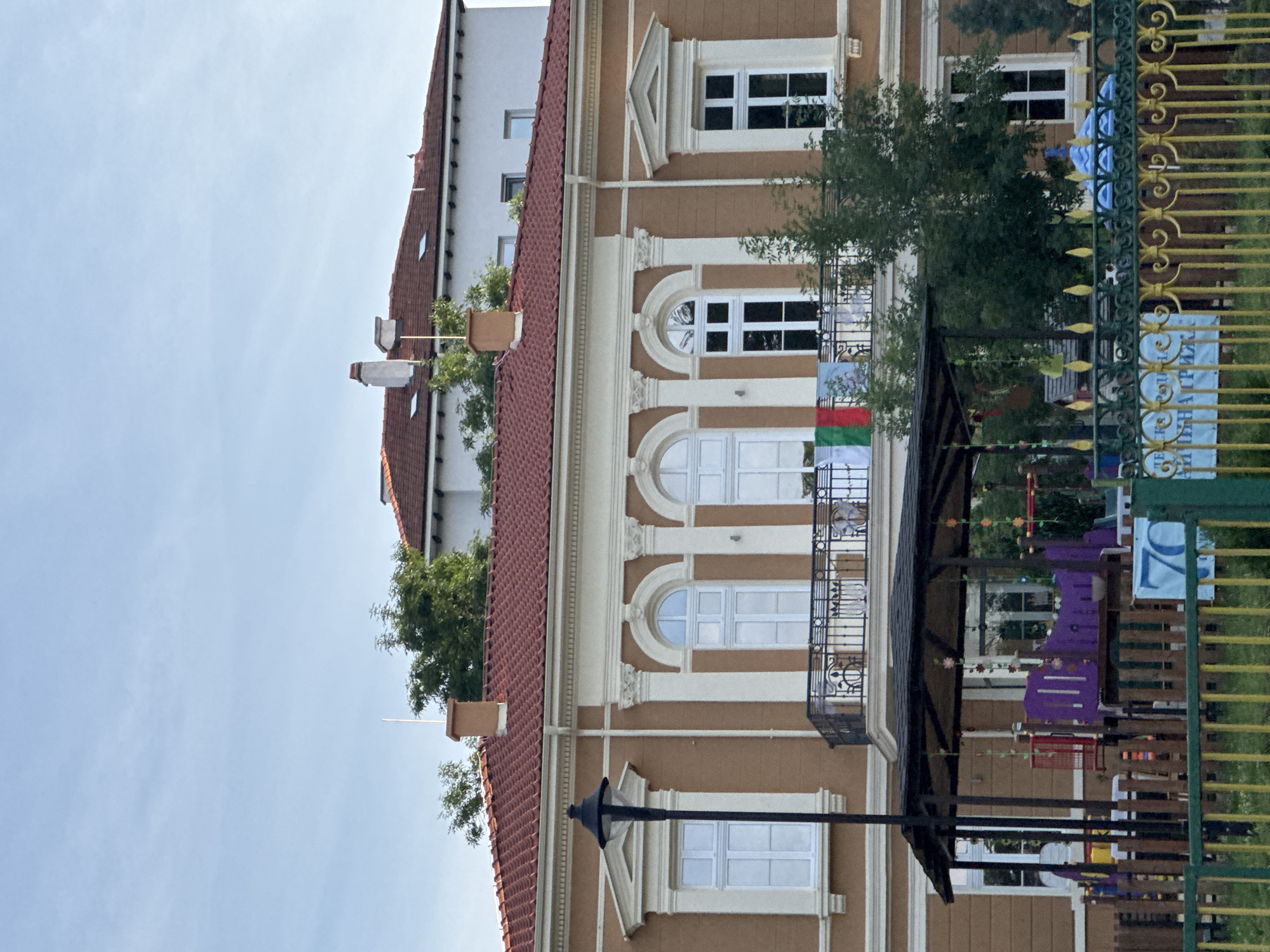 The image shows the facade of a building in Plovdiv, Bulgaria, with white-framed windows and light brown stucco detailing. A red and white tiled roof tops the building. A Bulgarian flag is visible on one of the balconies. A wrought-iron fence runs along the bottom of the image, and part of a children's playground is visible behind the fence. The sky is light blue and clear.