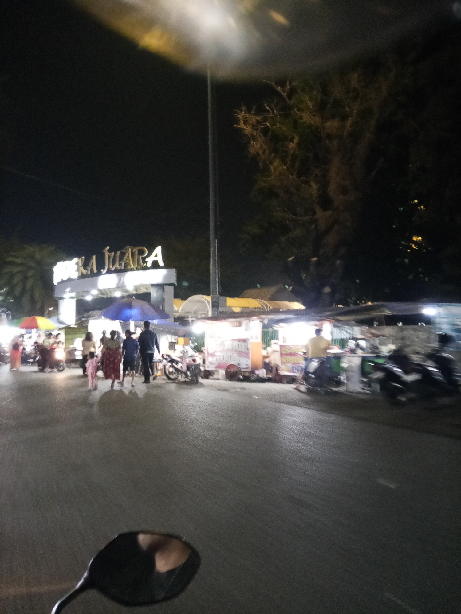 The image depicts a bustling night scene in front of a building with illuminated signage that reads "BUKA JUARA". Several people are walking and standing around, some gathered near food stalls. There are motorcycles parked and some are in motion, suggesting a busy atmosphere. The scene appears to be an outdoor market or food court. Based on the "BUKA JUARA" signage, this location is likely a culinary or entertainment spot in Lubuklinggau, Indonesia, as "Buka Juara" translates to "Open Champion" and often signifies a place known for good food or competitive events. The foreground shows a motorcycle's rearview mirror, indicating the photo was taken from a moving vehicle.