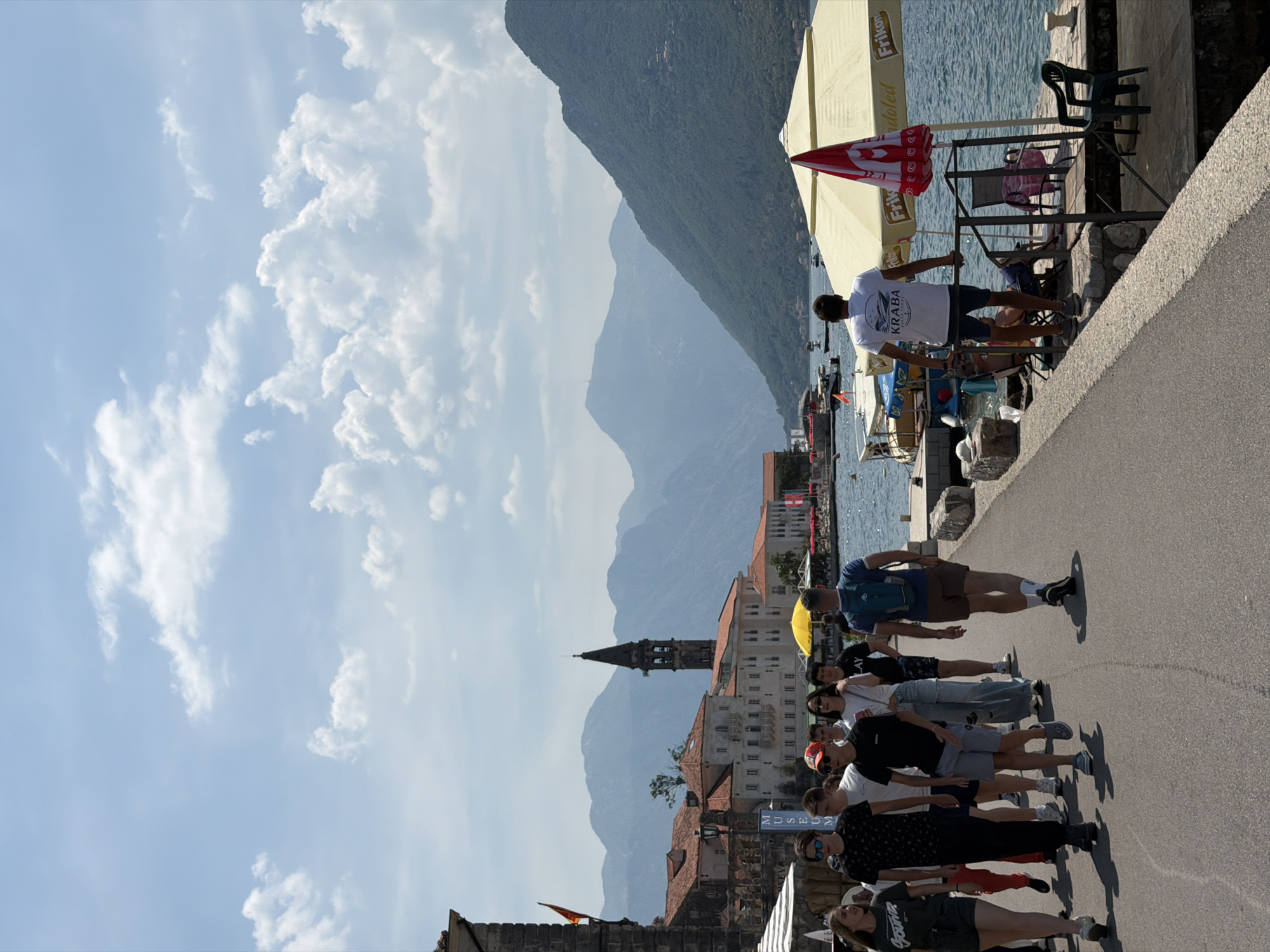 In the city of Perast, Montenegro, a group of people walks along a paved promenade beside a body of water. The promenade has a stone border on the water side. In the background, buildings with terracotta roofs and a prominent bell tower suggest historical architecture. Mountains rise behind the town, and a blue sky with scattered clouds provides the backdrop. The water is calm with several boats docked. Some individuals are walking towards the viewer, while others are facing away. One person stands facing the water, leaning on a railing. Umbrellas and seating are visible on a section of the promenade near the water.