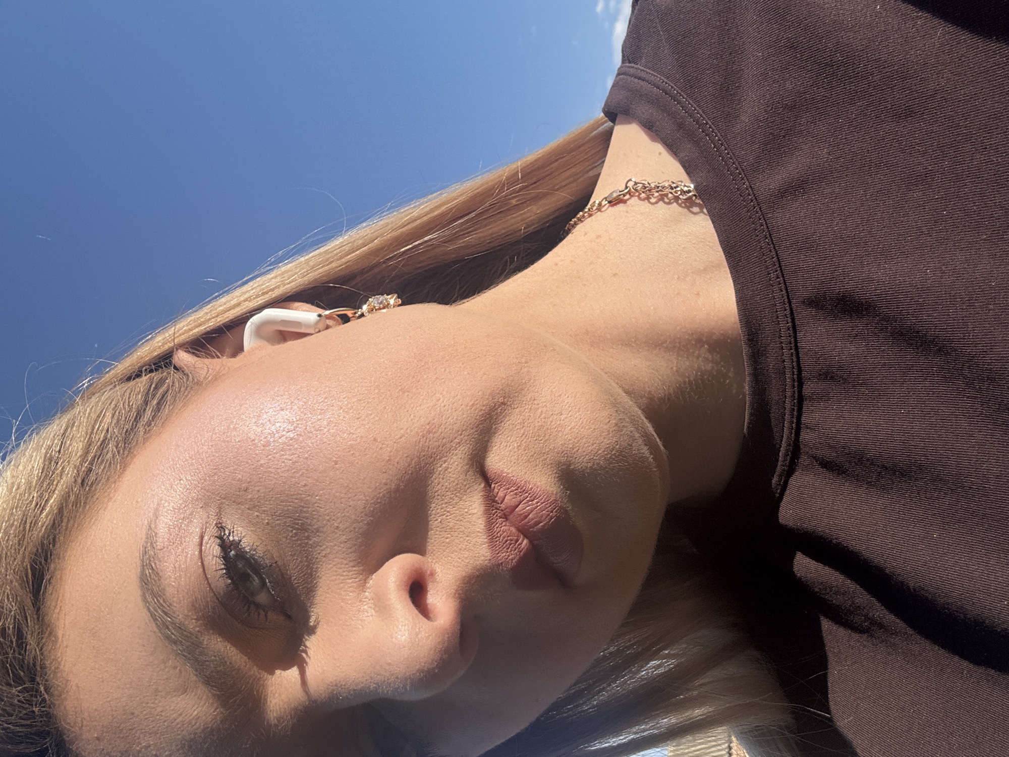 A close-up, low-angle outdoor shot features a woman's face and upper torso against a bright blue sky. She has light blonde hair and is wearing a dark brown, ribbed t-shirt. Her right ear is visible, adorned with a white earbud and a dangling gold earring with a clear stone. A gold chain necklace is also visible around her neck. Her skin appears to be slightly sun-kissed, with subtle makeup, including mascara and a nude lipstick. Sunlight casts a shadow across her forehead and cheek. The background is a clear, cloudless blue sky. No specific city or location details are discernible.