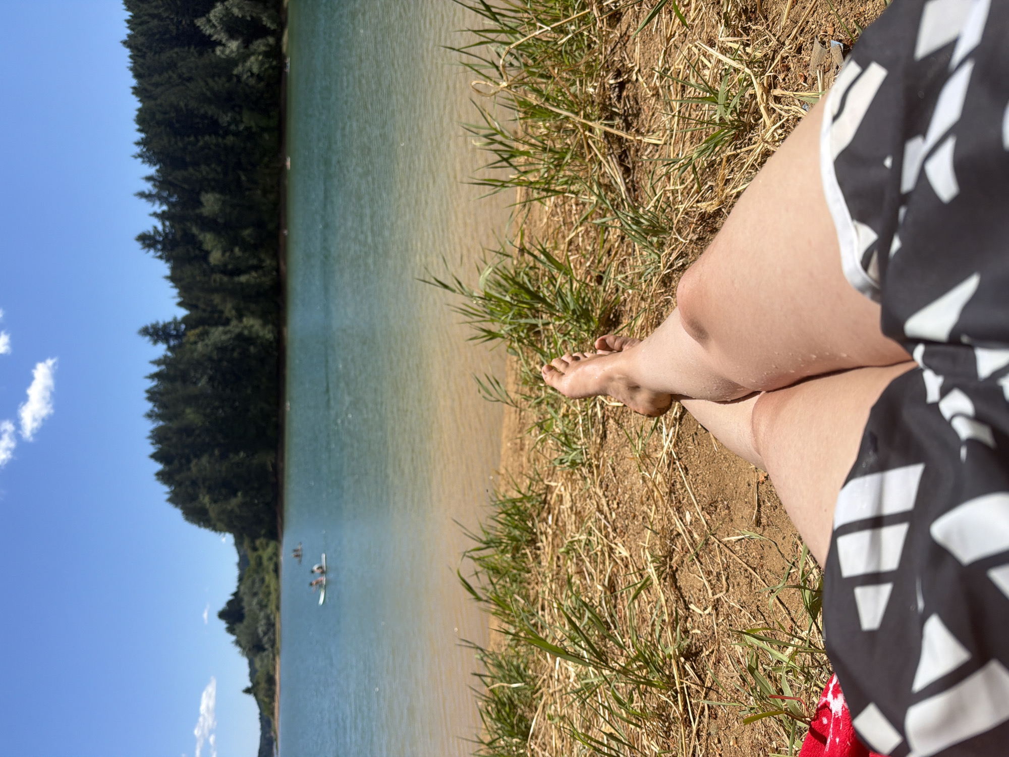 The image depicts a personal perspective of a person lying on the dry grass and sand near a body of water. The person is wearing black and white patterned shorts. Their legs and feet are visible, with one leg bent at the knee. The foreground is dominated by the dry grass and sand. In the mid-ground, a calm body of water extends towards a shoreline lined with a dense forest of evergreen trees. The water is a clear blue-green, and a few small boats with people on them are visible in the distance. The sky above is a bright blue with a few scattered white clouds. The scene suggests a warm, sunny day at a lake or reservoir. The location is not explicitly stated as Cherry Grove, United States in the image itself.