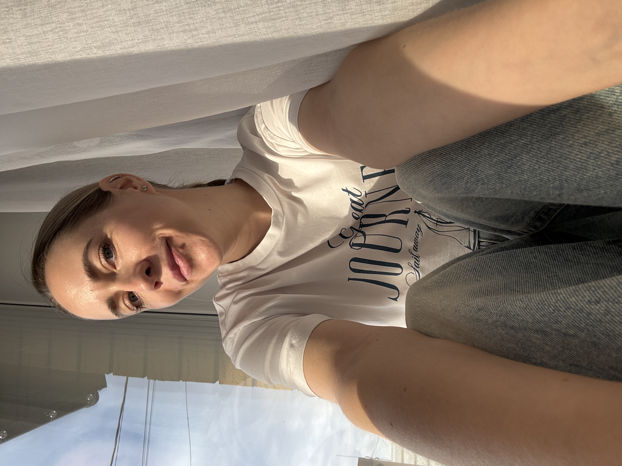 A selfie of a fair-skinned woman is displayed, taken from a low angle. She is facing the camera with a slight smile. She is wearing a white t-shirt with a navy blue print. She is also wearing denim jeans. The background consists of a window through which a cloudy sky is visible. A white curtain is partially visible in the upper part of the image. The location may be a domestic setting, but cannot be confirmed. There is no verifiable information about this being taken in the city of Chaikovsky, Russia.