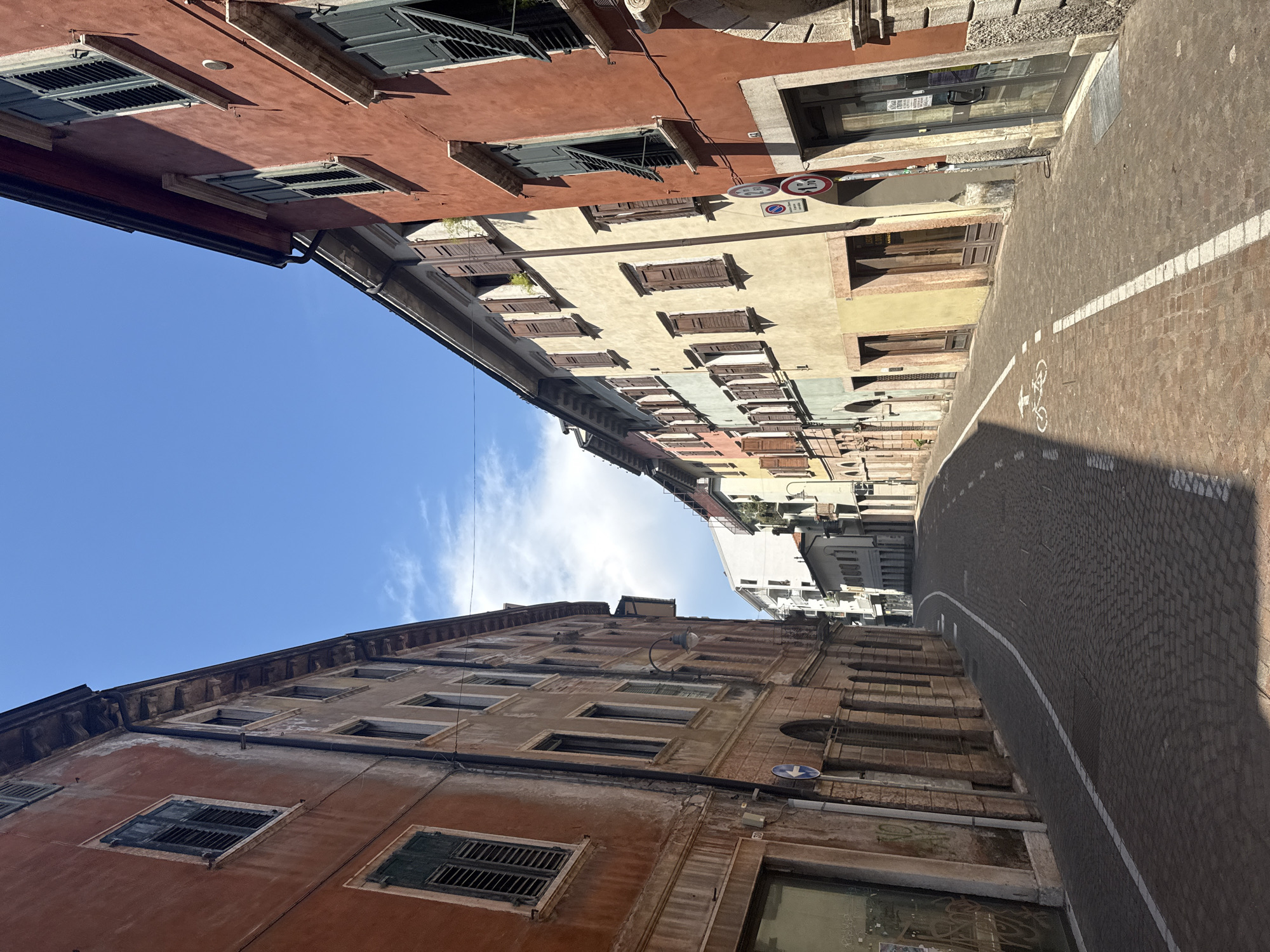 The image shows a street in Trento, Italy, with tall, colorful buildings lining both sides. The street is paved with cobblestones and has a bike lane marked by white lines. The buildings have traditional architecture with wooden shutters on the windows. The sky is blue with a few clouds. There are no people or animals visible in the image. The scene is quiet and appears to be from a sunny day.