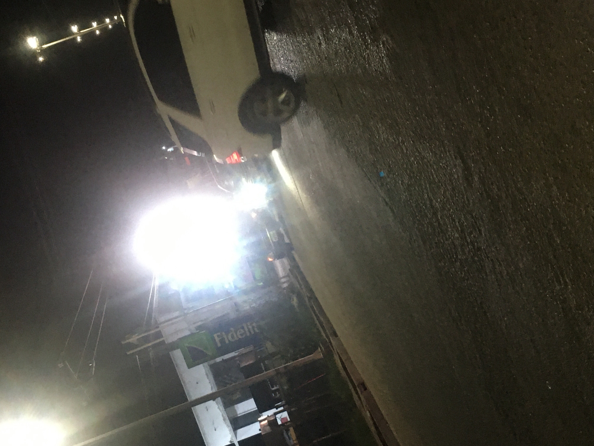 At night in Ado, Nigeria, the image shows a white car parked on a wet, dark street under a bright street light. Nearby is a building with a sign that includes the word "Fidelity". Other street lights are visible in the distance.