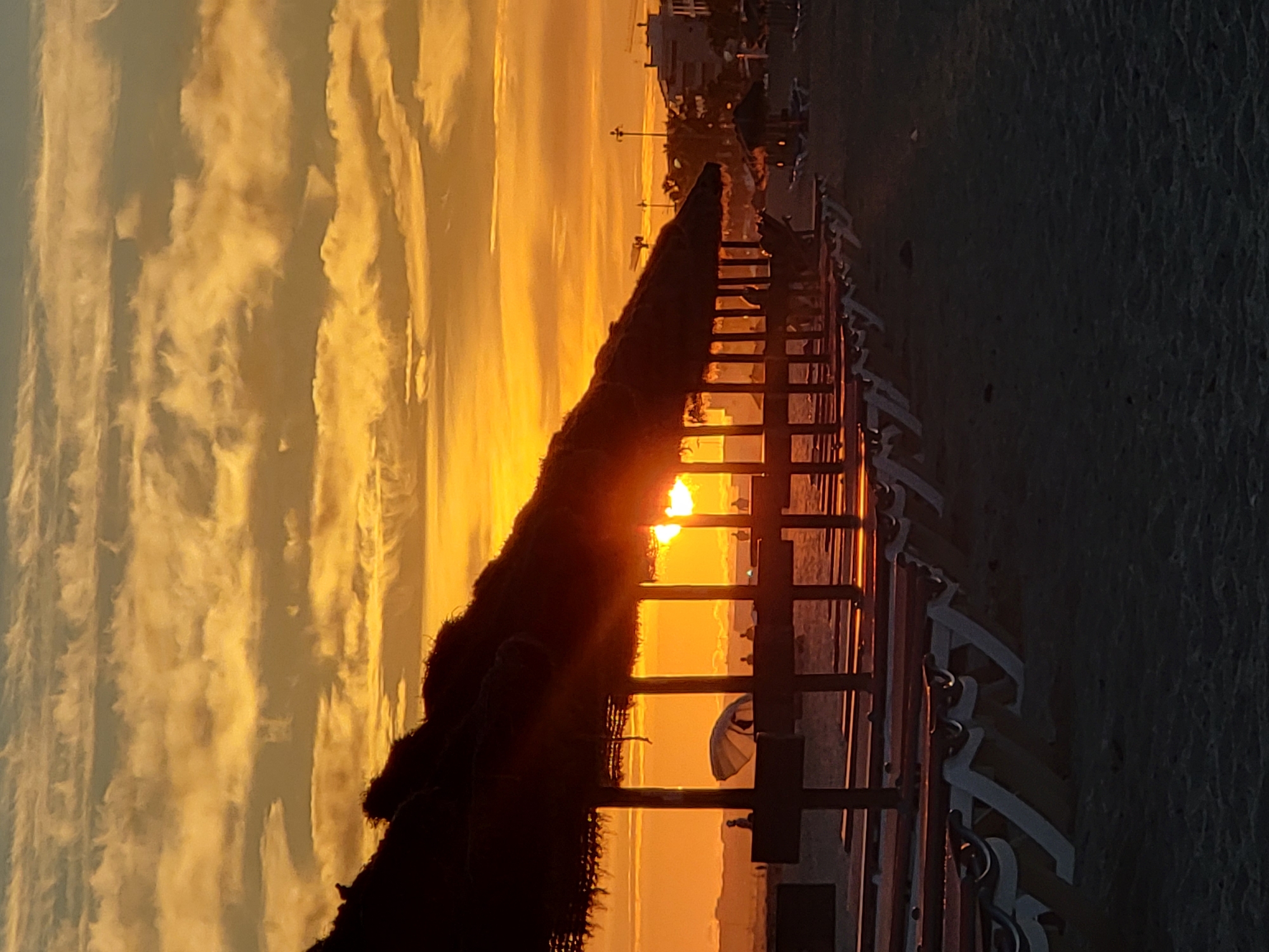 The image depicts a sunset scene on a beach in Quarteira, Portugal. In the foreground, a row of empty beach chairs with white frames and red accents are lined up on the sand, partially covered by thatched-roof beach umbrellas. The sun is setting in the background, casting a warm orange glow across the sky and clouds. The sun's rays are partially visible through the gaps between the umbrellas and their support structures. Silhouettes of people can be seen in the distance, as well as buildings and streetlights along the coastline. The overall atmosphere is tranquil, with the natural beauty of the sunset dominating the scene.