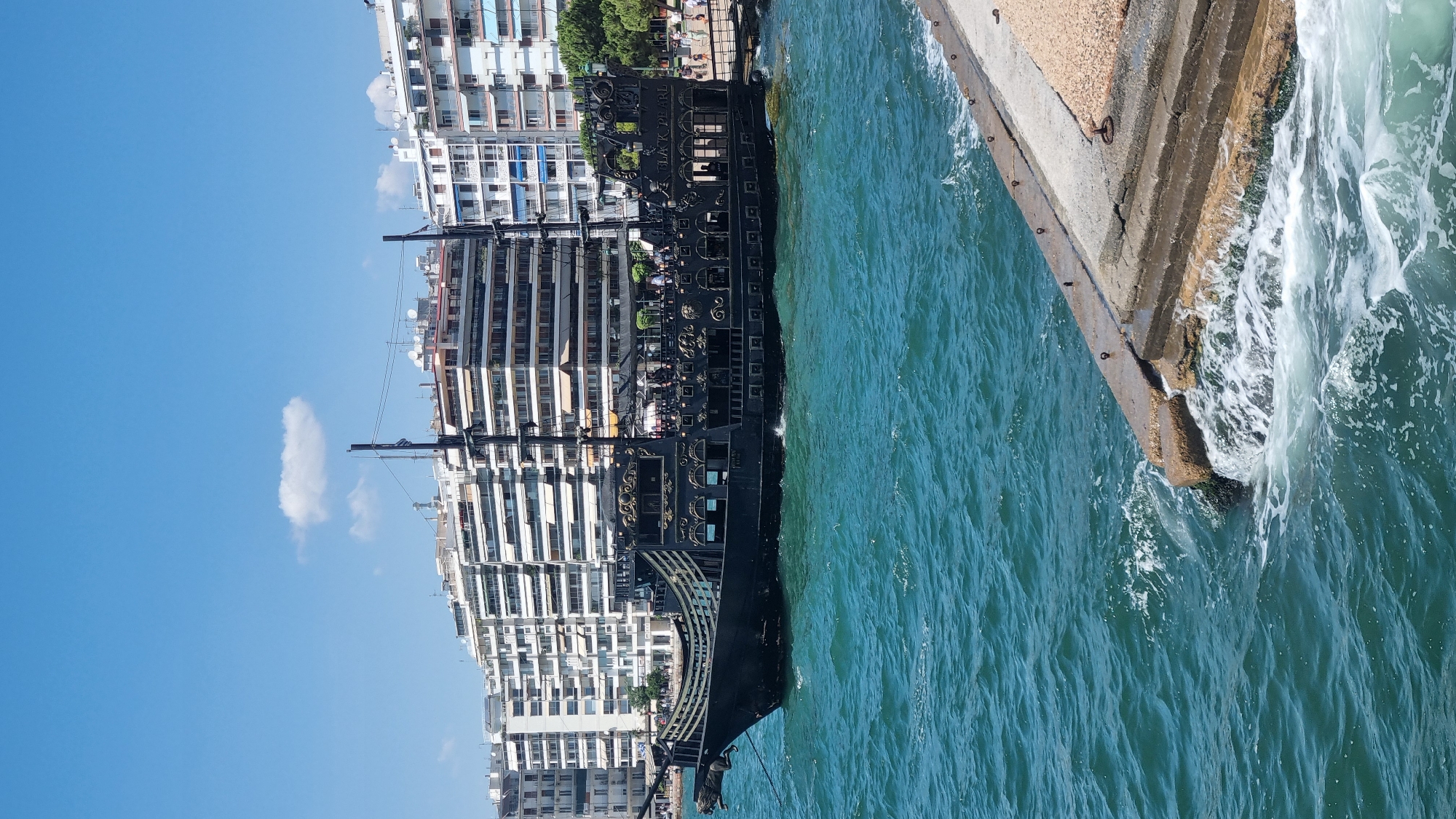 A black, elaborately decorated pirate ship named "Black Pearl" is docked in the turquoise waters of Thessaloniki, Greece, with a backdrop of modern apartment buildings lining the waterfront. The ship has a prominent bowsprit with a lion's head figurehead and several masts with rigging. People are visible on the deck of the ship, and some are standing on the pier. The water is choppy, with white foam created by waves lapping against a concrete pier in the foreground. The sky is a clear blue with a single, small white cloud. The scene depicts a bright, sunny day by the sea in an urban setting.