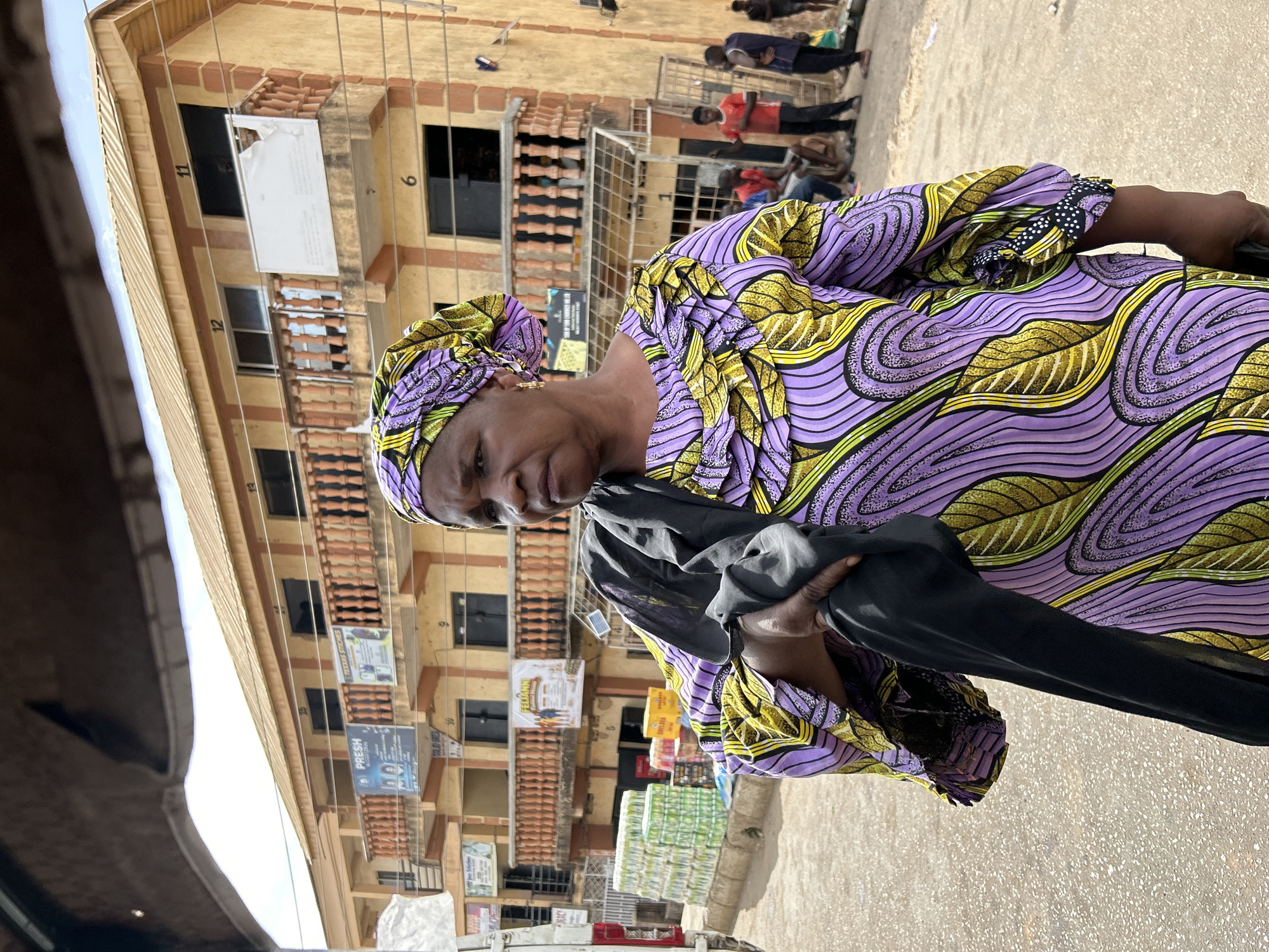The photo shows a woman in a purple and yellow patterned dress and headwrap, holding a black garment. The photo was taken in Jos, Nigeria. In the background is a multi-story building with various commercial advertisements on its facade. People are present in the background. Stacks of bottled drinks are stacked near the street. The street is paved.