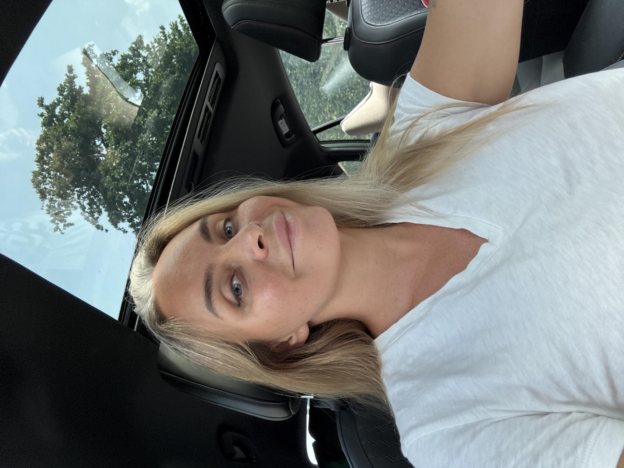A woman is seated inside a car, looking at the camera. She has blonde hair with gray streaks and is wearing a white V-neck t-shirt. The car has a sunroof open, revealing a glimpse of a tree and the sky. The interior of the car, including the headrests and seating, is visible. The image is taken from a front-facing perspective, capturing the woman's upper body and face. The scene appears to be outdoors, possibly on a sunny day, given the brightness and open sunroof. The location in Tambon Don Sak, Thailand, is not visually identifiable from the provided image.