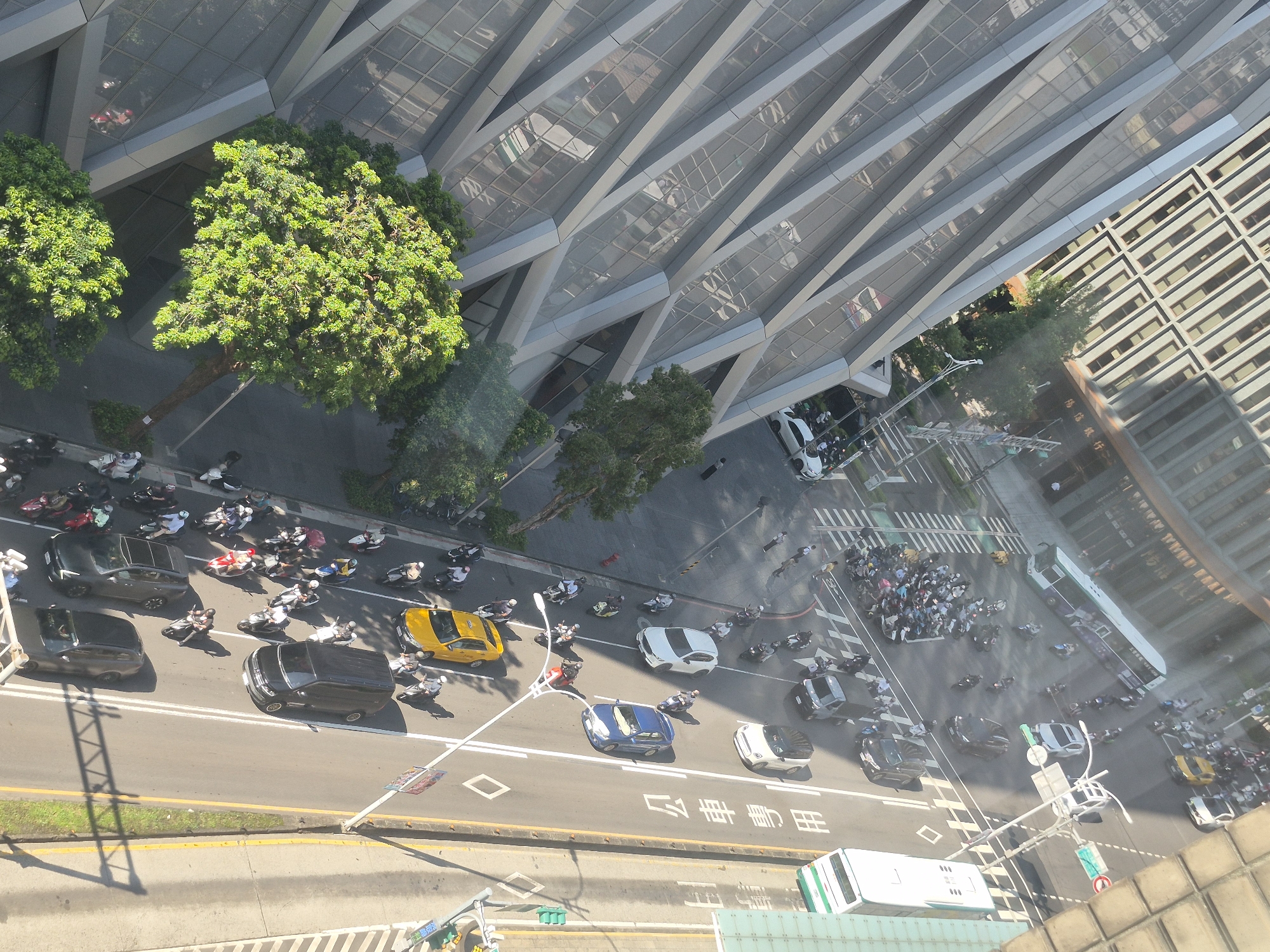 This aerial view captures a busy street scene in Taipei, Taiwan. The image shows a multi-lane road filled with numerous motorcycles, cars, and a bus. The vehicles are in motion, navigating the intersection, with many motorcycles clustered together. Pedestrians are also visible, some crossing the street, others standing on the sidewalks. The surrounding environment includes modern architecture, characterized by a building with a distinctive geometric facade, and lush green trees planted along the roadside. The lighting suggests it is daytime, with shadows cast by the structures and vehicles. The overall impression is one of urban activity and traffic flow in a developed city.