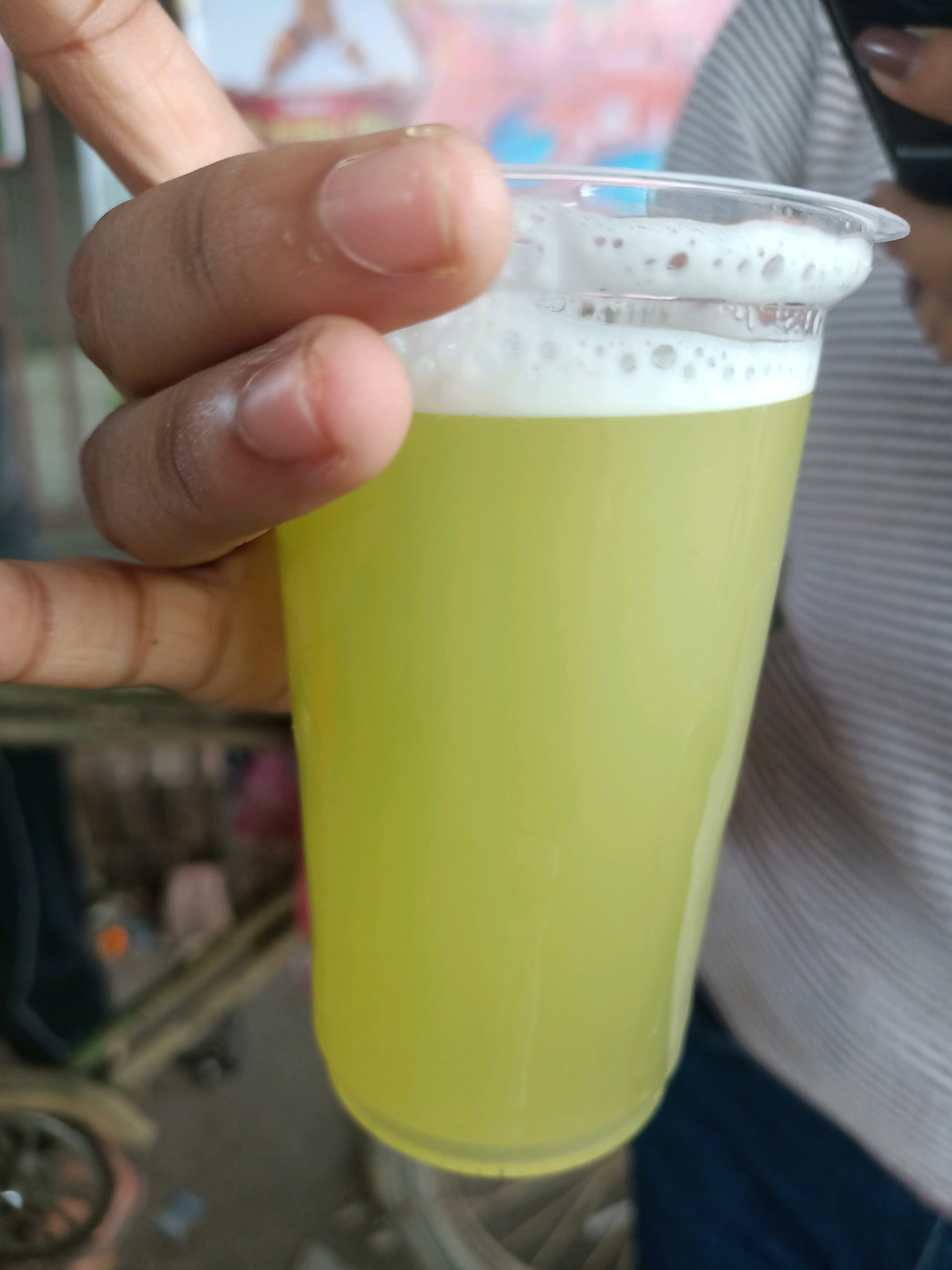 A person's hand, with visible fingernails and knuckles, is holding a clear plastic cup filled with a light yellow, frothy liquid. The cup is filled to the brim, with a layer of white foam on top. In the background, a person in a gray and white striped shirt is partially visible, holding a dark object to their ear, possibly a phone. The environment appears to be outdoors, with a blurred background suggesting a street or market scene in Barrackpore, India, indicated by the presence of what appears to be bamboo scaffolding and other indistinct objects.