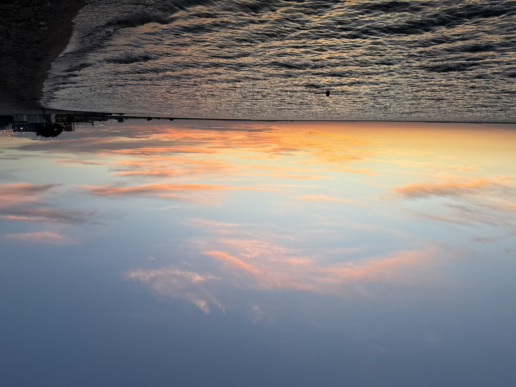 The image depicts a tranquil scene at the shore of a body of water, likely during sunset. The sky is painted with hues of orange, pink, and blue, with wispy clouds scattered across it. The water is calm, with gentle ripples reflecting the colors of the sky. A solitary person is visible in the water, swimming or floating. The shoreline on the right is a dark sandy beach, with some distant buildings and streetlights indicating a developed area. The overall impression is one of peace and natural beauty. Without further information, pinpointing the exact location within Kokshetau, Kazakhstan, is not possible, but it is consistent with a lakeside or riverfront setting.