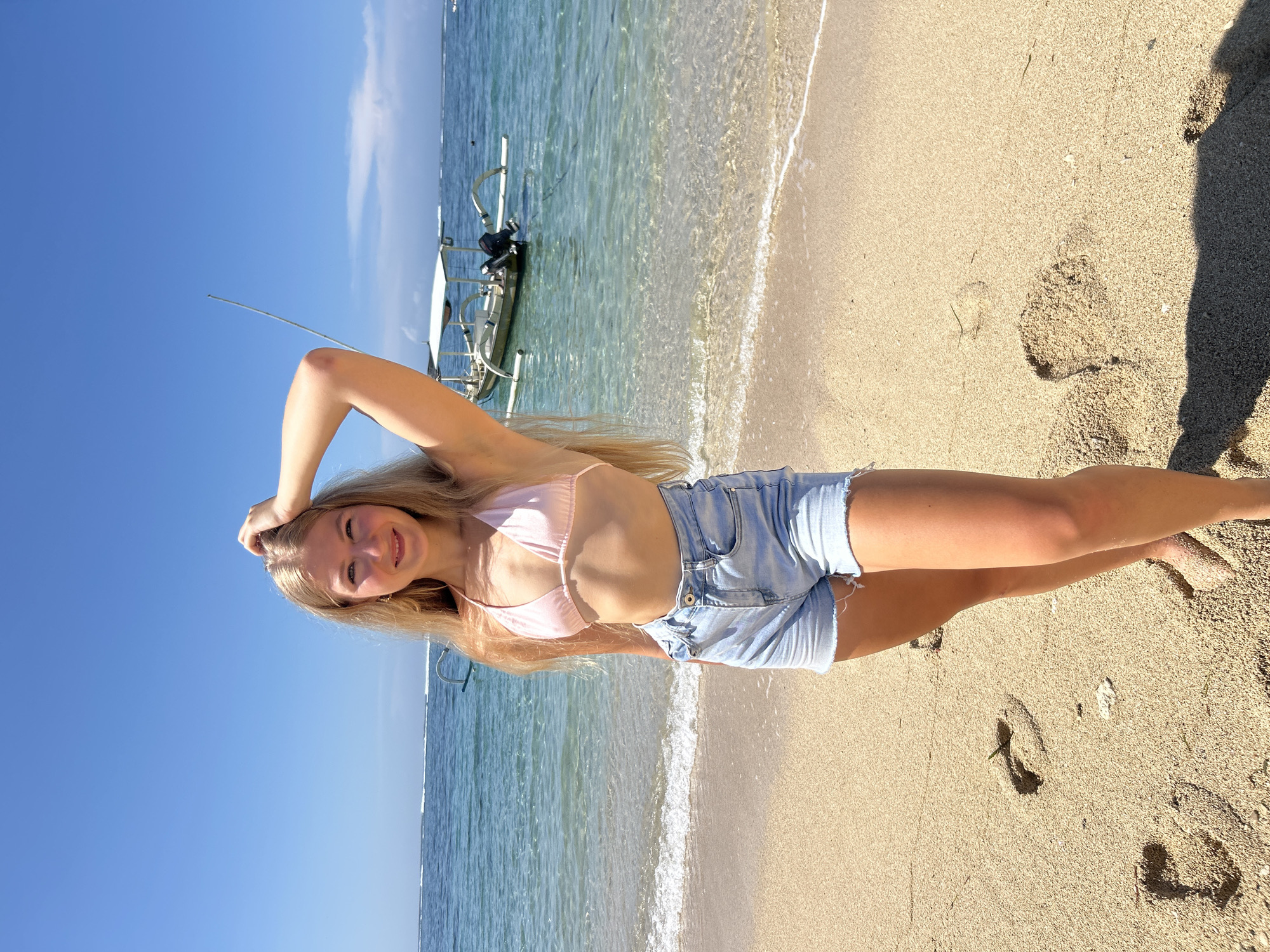 A young blonde woman in a pink bikini top and denim shorts is standing on a sandy beach with a boat in the background, under a bright blue sky and sunny conditions. The location is likely in or around the city of Kuta Selatan, Indonesia, based on the common location of boats on tropical beaches there. The woman has one arm raised to touch her head and is facing the camera with a slight smile. The beach has visible footprints in the sand, and the water is clear with small waves.