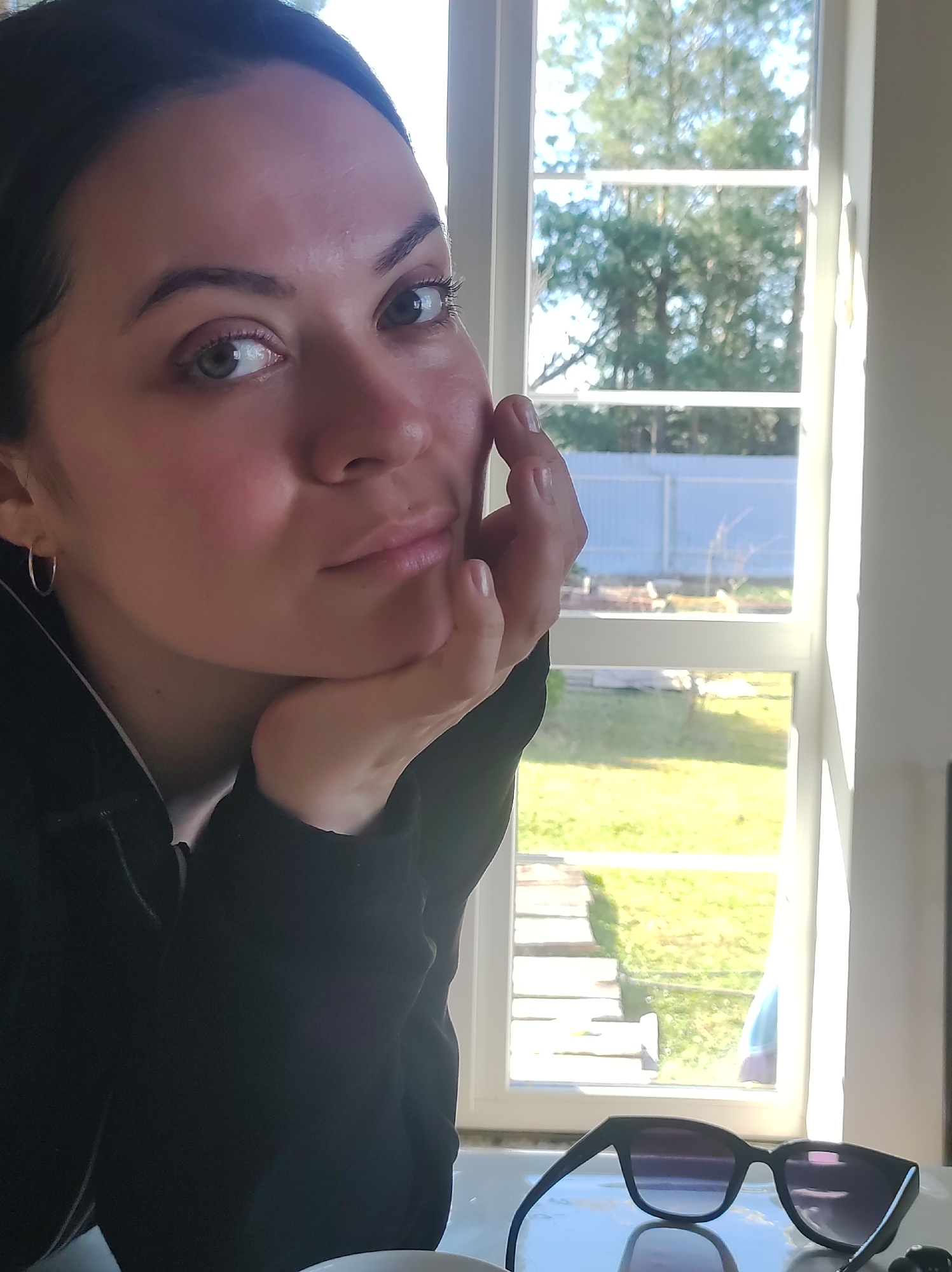 A woman with dark hair and fair skin is sitting indoors with her head resting on her hand, looking towards the camera. She is wearing a dark colored hoodie with a white lining around the neckline. A pair of sunglasses is lying on a surface below. Behind her is a white-framed window providing a view of a yard with green grass, some trees and a white fence in the distance. The location cannot be identified as Фоки, Russia from the image alone.