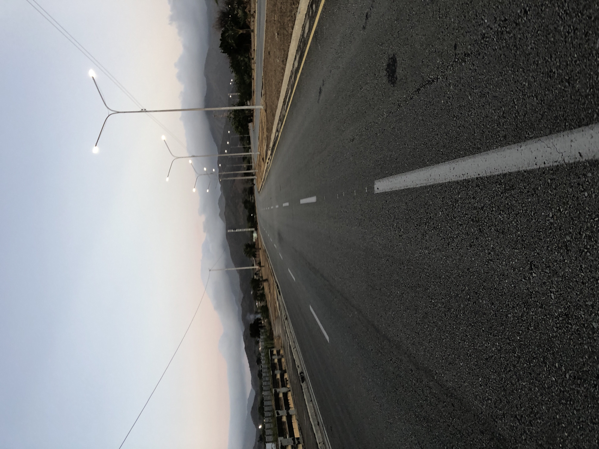 The image shows a straight stretch of asphalt road, possibly in Altagracia, Venezuela given the prompt, with white painted lane markings. The road is flanked by a curb and a narrow strip of land. A series of streetlights line the left side of the road, extending into the distance. Beyond the streetlights, mountains are visible in the background, partially obscured by haze or low clouds. The sky is overcast and appears to be at dusk. The asphalt appears new or well-maintained, with visible aggregate texture. There are no people or other objects immediately visible on the road.
