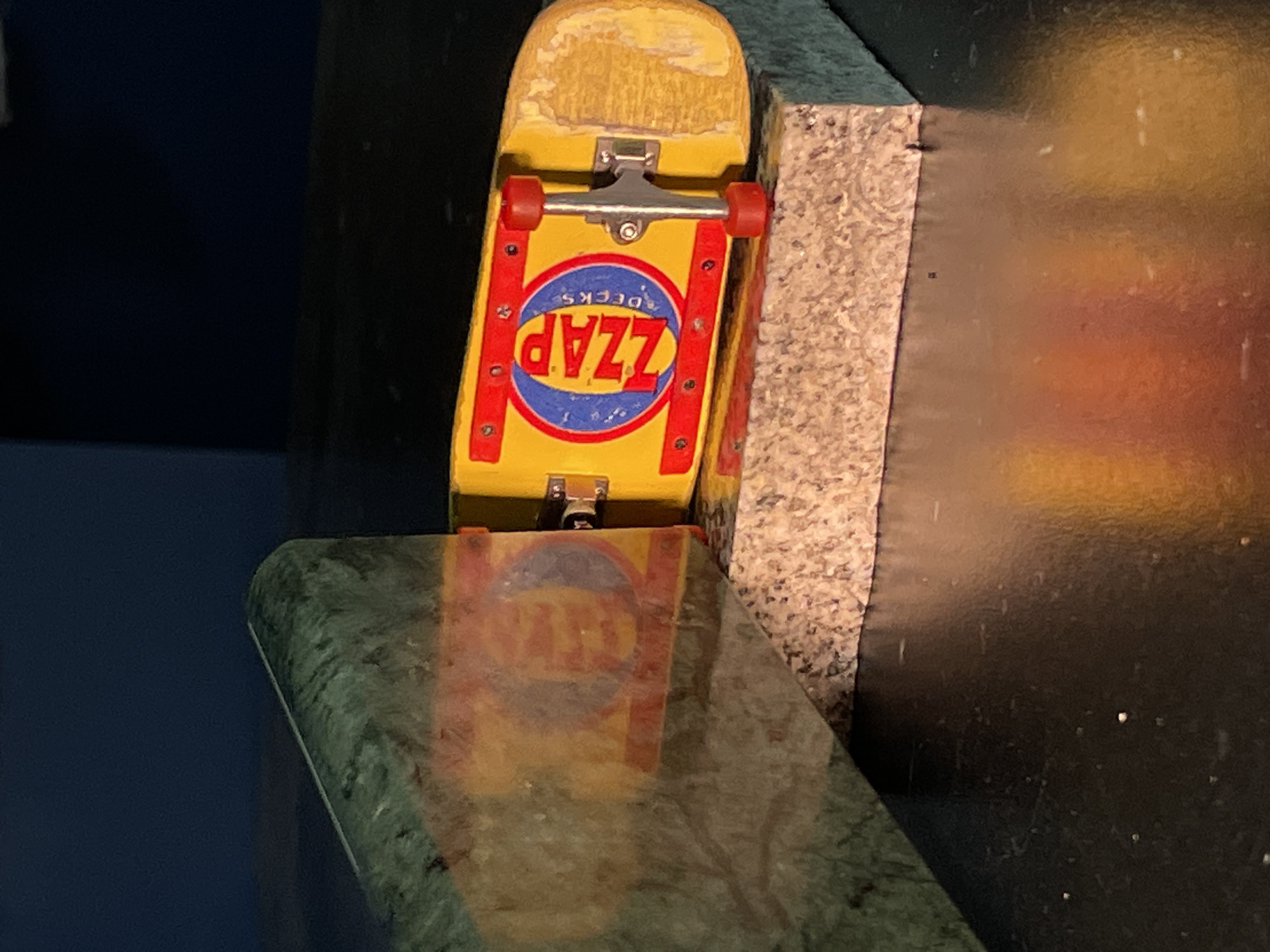 A yellow Zzap brand fingerboard is leaning against a light gray speckled object and a dark object. The fingerboard has red rails and red wheels. The fingerboard is reflecting on a surface below it. The environment appears to be indoors. There is no evidence to suggest a location in Yekaterinburg, Russia.