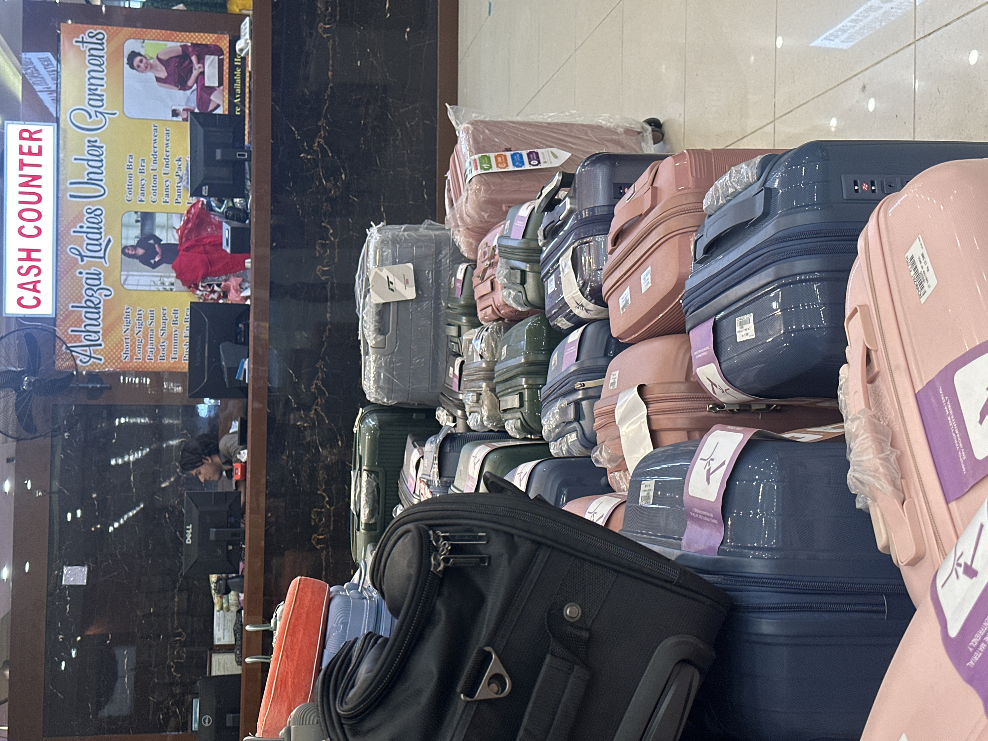 The scene depicts a retail environment, likely a shop, located in Quetta City Tehsil, Pakistan. The focal point is a stacked display of suitcases in various colors and sizes, including pink, blue, gray, green, and black. The suitcases appear to be new and packaged, some bearing labels and protective wrapping.
In the background, a "Cash Counter" sign and advertisement for "Achakzai Ladies Under Garments" is visible, suggesting the store sells women's clothing and related items. The ad lists various garment types, including nightwear, lingerie, and shapewear.
A person is seen sitting at a computer behind the counter, possibly a store employee. A Dell monitor is visible. The environment suggests an indoor commercial setting, possibly a department store or a specialized luggage shop.