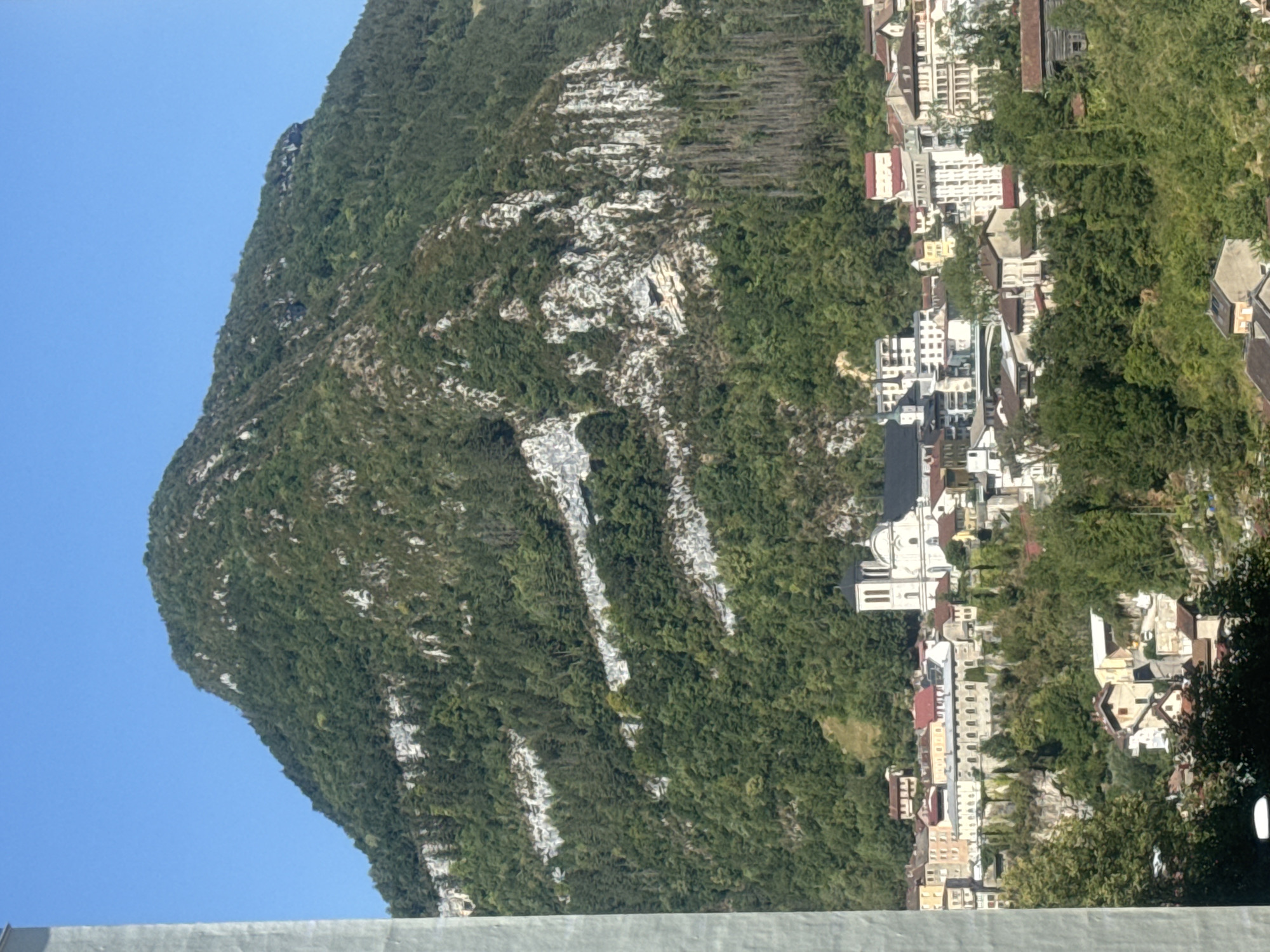 The image shows a view of the city of Saint-Claude, France, nestled at the foot of a large, green mountain. The mountain is covered in dense forest, with several visible white rock strata formations running horizontally across its face. A small town is situated on the slopes leading up to the mountain, characterized by a mix of traditional buildings with red tile roofs and more modern structures. A prominent white church with two towers stands out in the center of the town. The sky above is clear and blue. The foreground of the image features trees, and a portion of a grey, vertical structure is visible on the far left edge of the frame.