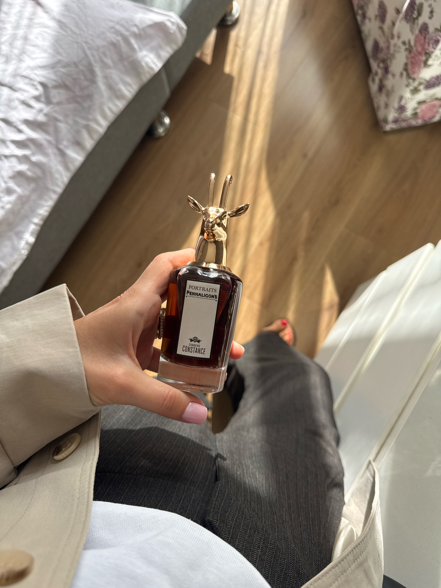 A person's hand, with light pink nail polish, holds a clear glass perfume bottle with a dark amber liquid and a distinctive gold stag head stopper. The bottle has a white label that reads "PORTRAITS PENHALIGON'S" and "CHANGING CONSTANCE". The person is wearing a tan trench coat over dark grey pinstriped pants and a white top. They are sitting on a white chair, and their feet are visible, with red nail polish on their toenails. The background consists of a wooden floor and a white draped object. The scene is illuminated by natural light casting shadows on the floor. The location is identified as Tbilisi, Georgia.