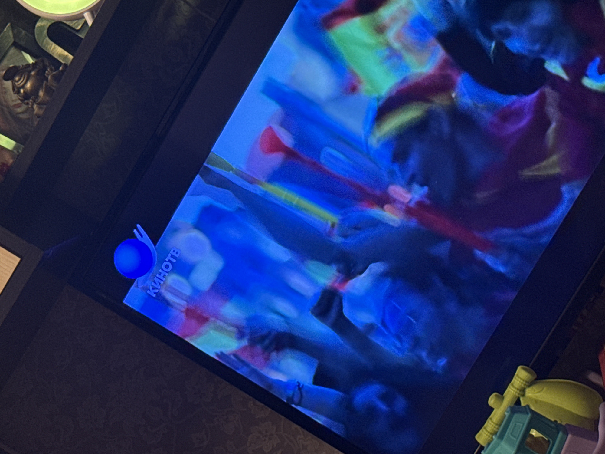 The image shows a television screen displaying an abstract, colorful image. The word "КИНОТВ" (KINOTV), which means "Cinema TV" in Russian, is visible on the screen. To the left of the screen, partially visible, is a decorative object, possibly a statuette, and a lamp. To the right of the screen are children's toys. The environment seems to be an indoor setting, likely a living room. Moscow, Russia is not discernible in this image.
