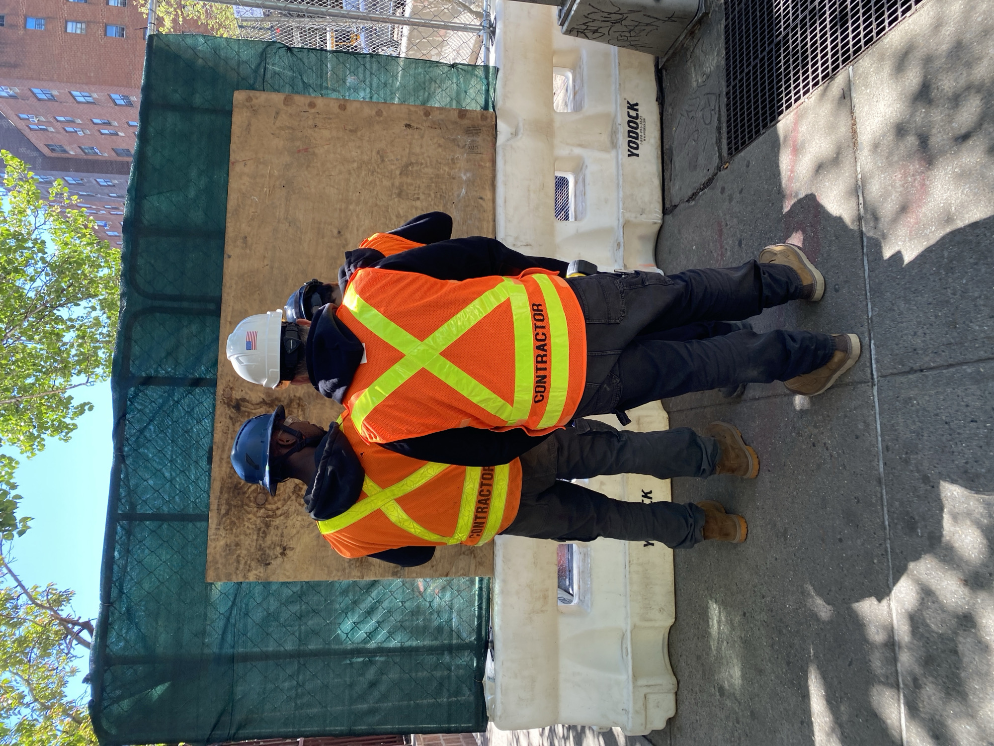 Two workers stand on a sidewalk in New York City, wearing orange safety vests labeled "CONTRACTOR." The first worker wears a blue hardhat, while the second has a white hardhat with a United States flag. The sidewalk is adjacent to a construction site protected by a green fence, a plywood panel, and white plastic barriers with the "YODOCK" brand name. A brick building is visible in the background, and foliage from trees is visible to the left. The ground is shaded, with sunlight hitting portions of the sidewalk. A metal grate is near the barriers.