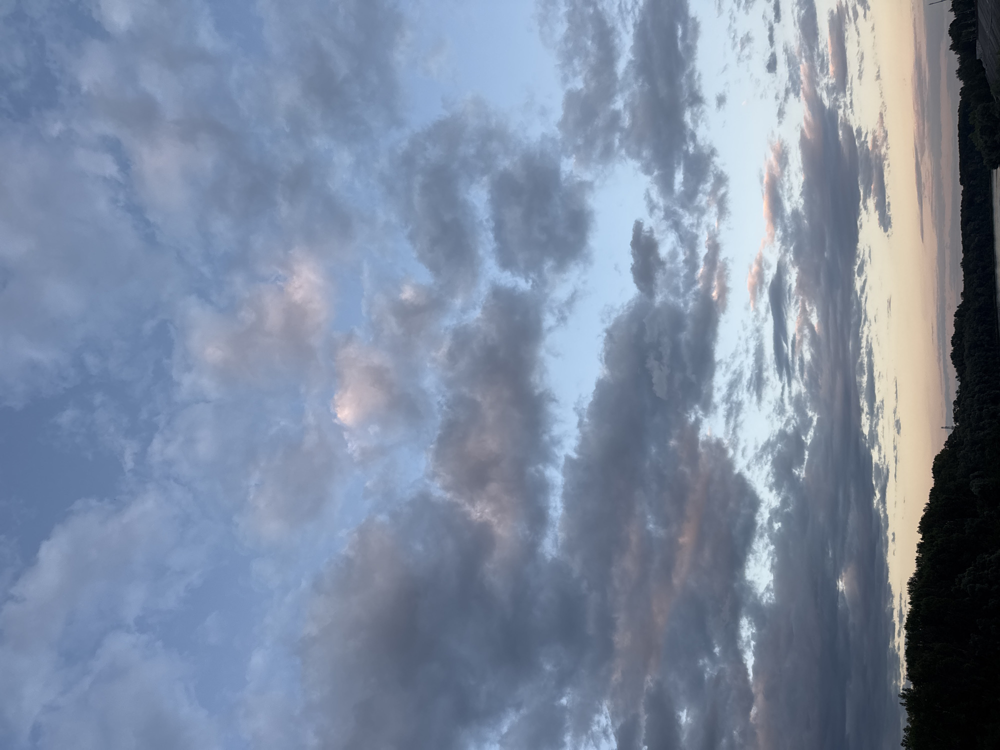 The image depicts a cloudy sky over a wooded area and a body of water, likely a river or lake, in Ufa, Russia. The sky is filled with a mix of dark gray and lighter, wispy clouds, with hints of pink and orange suggesting either sunrise or sunset. The tree line at the bottom of the image is dark and silhouetted against the lighter sky. A faint glow on the horizon indicates the sun is low. The scene is natural and serene, with no visible human activity. The overall impression is one of a peaceful natural landscape at dawn or dusk.