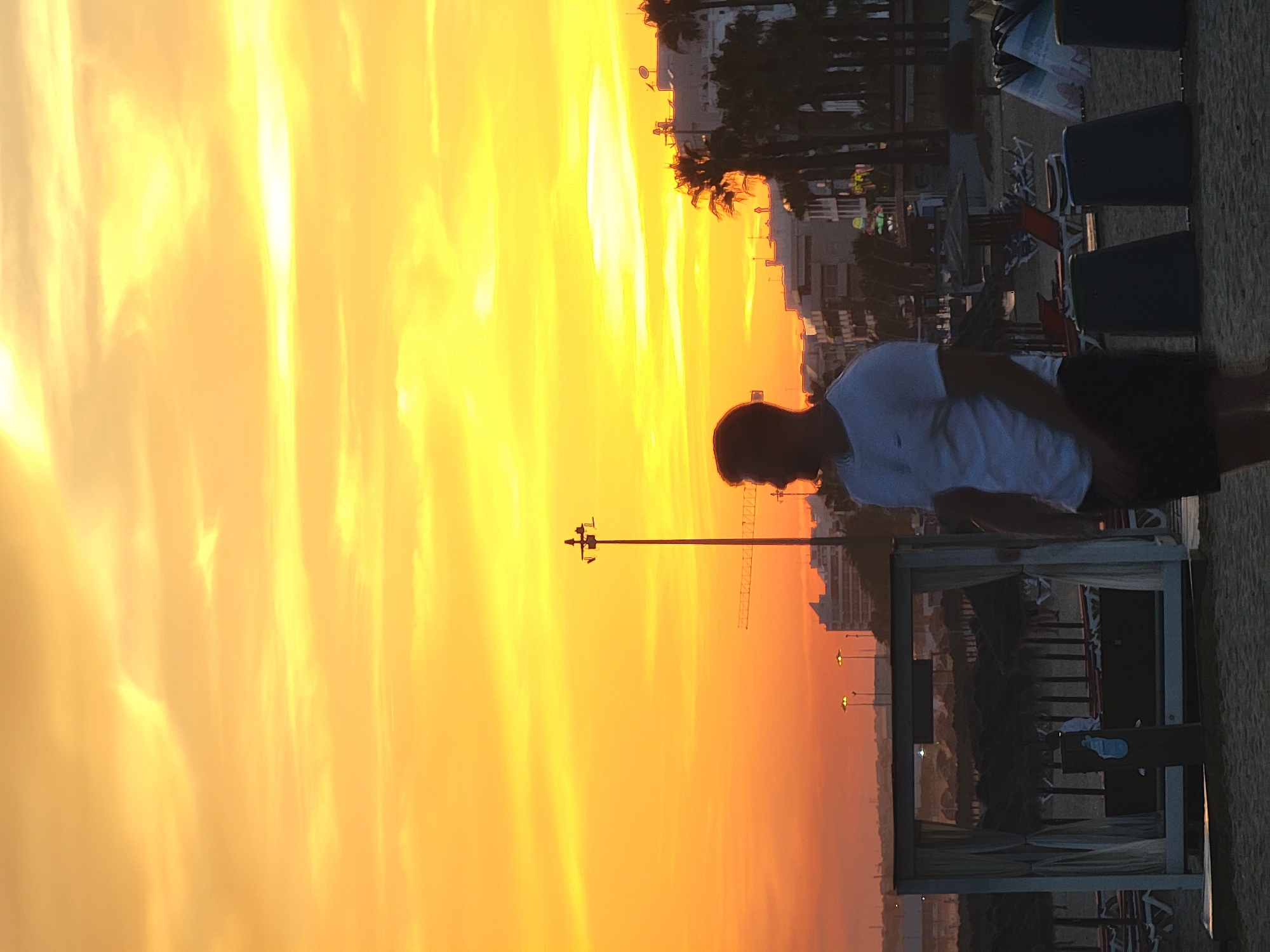 A full-shot, vertical, outdoor, low-angle, backlit photograph captures a man standing on a sandy beach in Quarteira, Portugal, during a vibrant sunset. The sky is a dominant feature, ablaze with intense shades of yellow and orange, with wispy clouds adding texture and depth. In the mid-ground, a silhouette of buildings, palm trees, and beach umbrellas lines the horizon, suggesting a coastal resort town. A lamppost stands prominently to the right of the frame, partially silhouetting a construction crane in the distance. The man, in the foreground on the right, is a dark silhouette wearing a white t-shirt and dark shorts, facing towards the left side of the image, away from the viewer. His posture is relaxed, with one hand in his pocket. To the left, a white cabana-style beach structure is visible, with drawn curtains. The overall atmosphere is serene and warm, characteristic of a summer evening by the sea.
