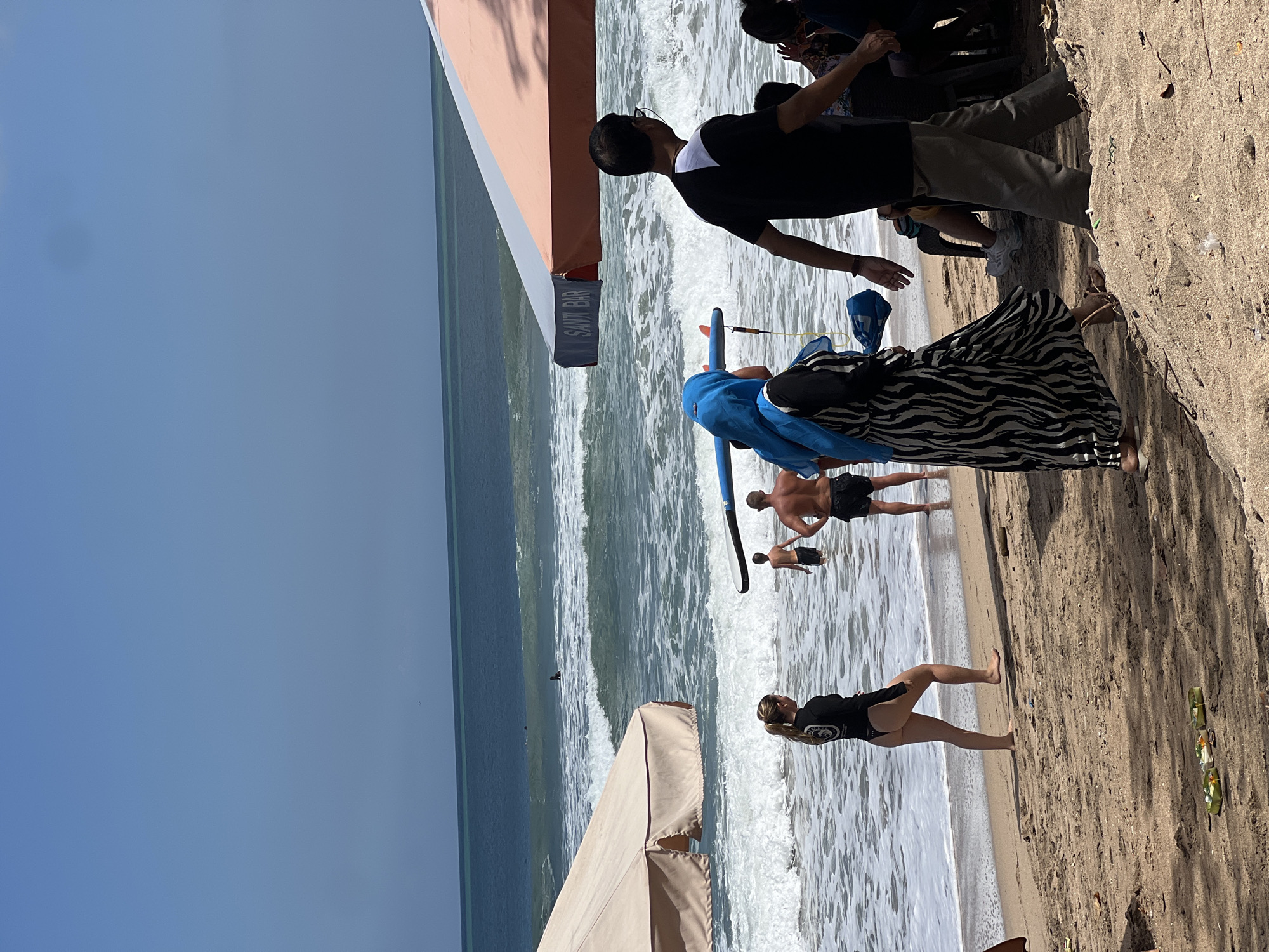 In the city of Kuta, Indonesia, a busy beach scene unfolds under a bright blue sky with a few scattered clouds. Waves crash onto the sandy shore, where several people are present. In the foreground, a woman in a black one-piece swimsuit walks along the wet sand. Further back, two men are standing in the shallow water, one with his back to the camera, and another with his arms by his side. In the mid-ground, a person wearing a blue hijab and a black and white zebra-striped dress walks with a surfboard. They are near a large beach umbrella that provides shade. To the right, a man in a black and white t-shirt and khaki pants walks away from the viewer, and other people are seated under another beach umbrella. The ocean stretches out to the horizon, with smaller waves breaking further offshore and a few surfers visible in the distance. The sand is scattered with debris, and small offerings of flowers and food are placed on the beach.