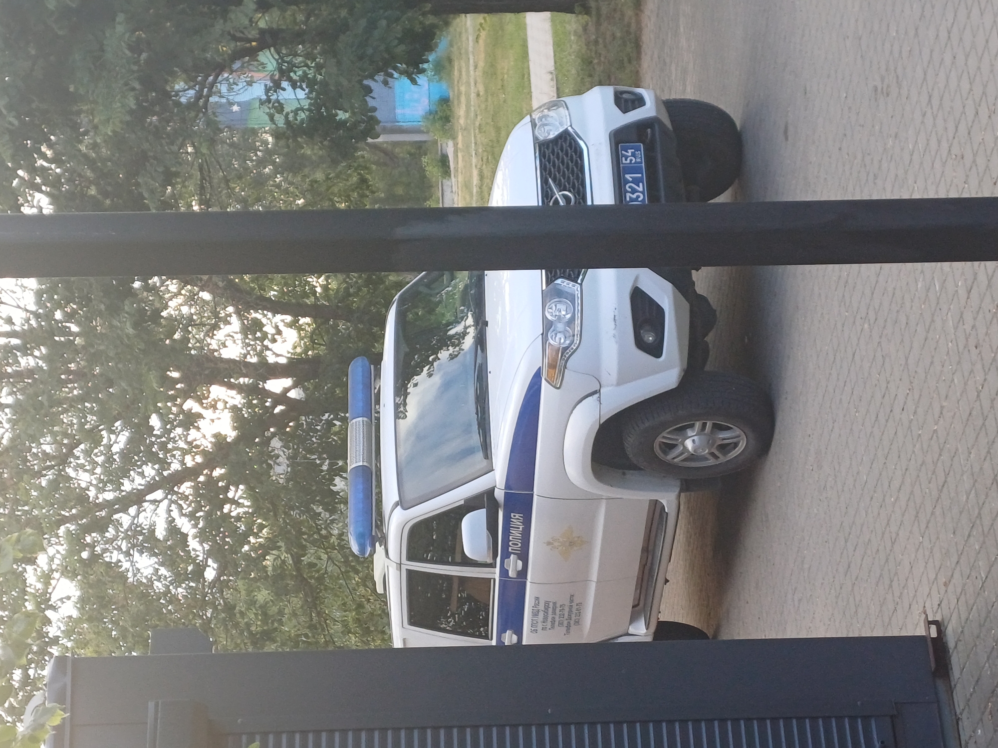 A white police SUV with blue and white markings is parked on a paved surface. The vehicle has a blue light bar on its roof. Text on the side of the vehicle is partially visible, indicating it is a police vehicle from Novosibirsk, Russia. The license plate reads "1321 54 RUS". Trees and a building are visible in the background. A dark vertical post is in the foreground, partially obscuring the vehicle.