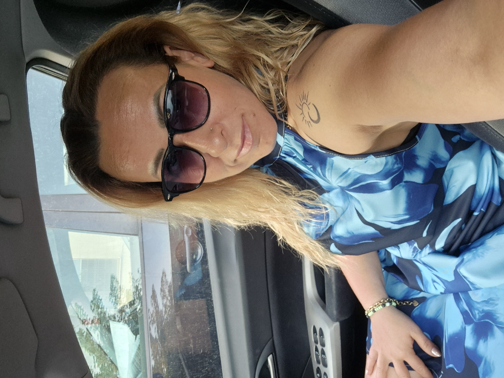 A blonde woman with sunglasses on, wearing a blue floral dress, is sitting in the driver's seat of a car. She has a tattoo of a sun on her left shoulder and is wearing a bracelet. The car's interior is visible, including the door panel with window controls and the passenger seat. The background shows a blurry reflection of trees and a building in the car window, suggesting an urban environment.