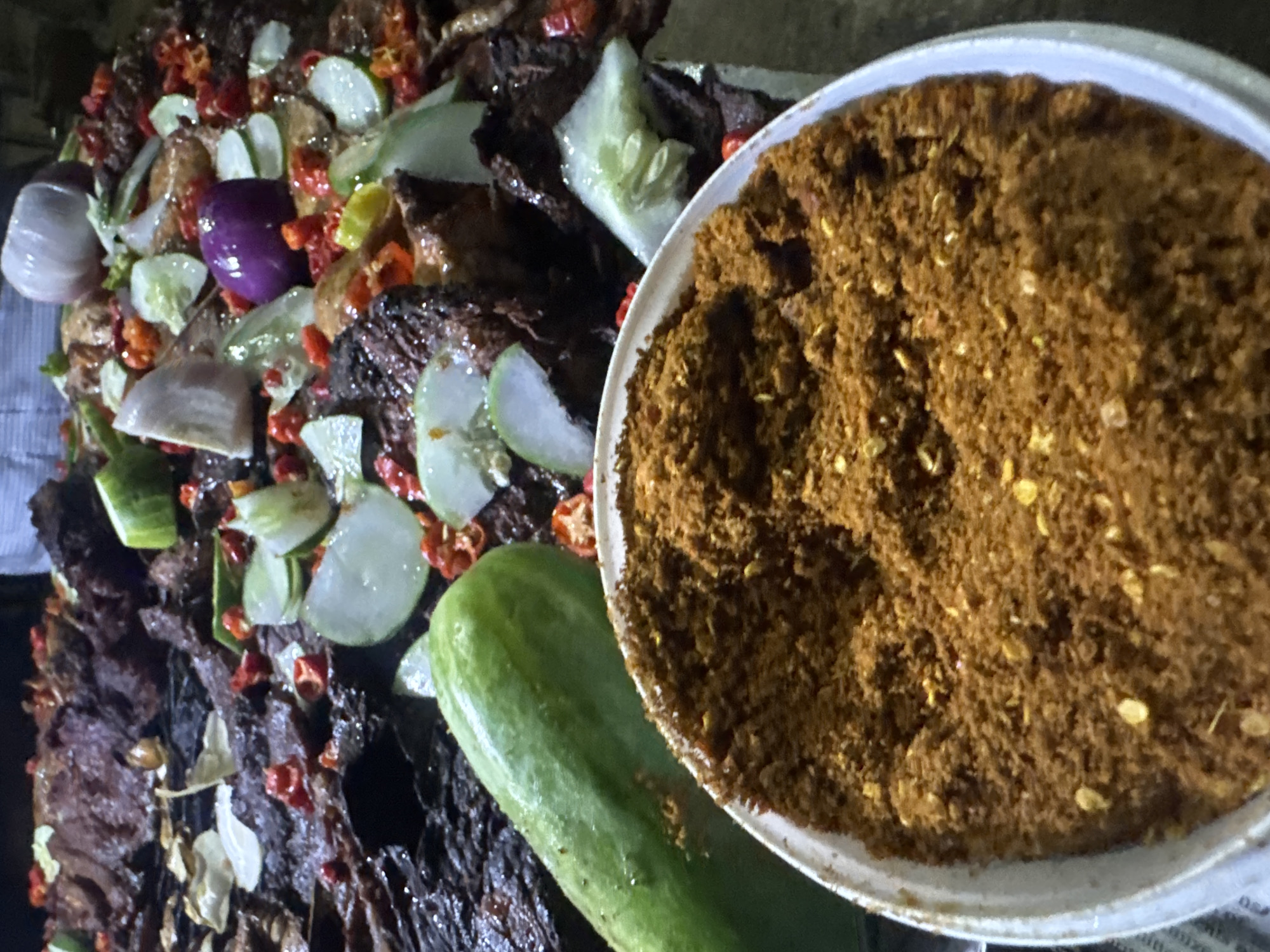 This media file captures a close-up view of a dish prepared with what appears to be dried meat, possibly a Nigerian delicacy like "Suya" or jerky. The meat is dark brown and textured, mixed with an assortment of ingredients. Among these are slices of cucumber, red chilies, and purple onions, adding color and freshness to the dish. A whole, green cucumber lies prominently in the foreground. To the right, a white bowl is filled with a generous portion of a reddish-brown spice blend, likely a seasoning mix for the meat. The overall presentation suggests a prepared meal or ingredients ready for consumption, common in a market setting or a home kitchen. The lighting is somewhat dim, possibly indicating an indoor setting or evening time, and there are no visible cues for weather. There is no discernible text in the image. The scene is focused entirely on the food, highlighting its ingredients and preparation. The context of Abuja, Nigeria, suggests this could be a representation of local cuisine.