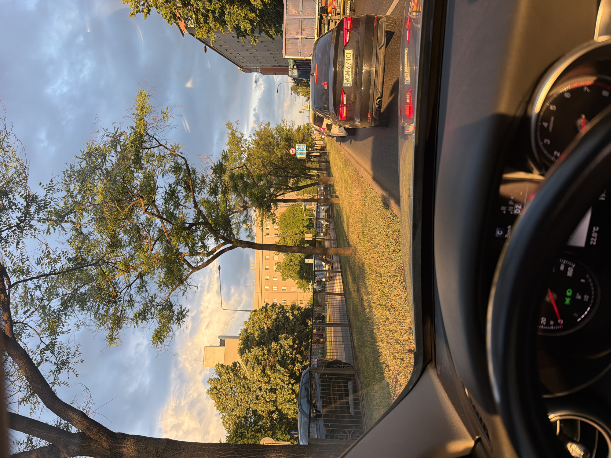 Captured from inside a vehicle, the image presents a street scene in what appears to be an urban area. The vehicle's dashboard and steering wheel partially frame the view, with the speedometer indicating a low speed or a stationary state. Outside, a dark-colored sedan with license plate "WGM 6710U" is visible ahead in the lane, followed by a SCANIA vehicle. To the side, a grassy strip separates the road from a pedestrian pathway lined with trees and a low fence. Buildings can be seen in the background under a partly cloudy sky, suggesting a late afternoon or early evening light.