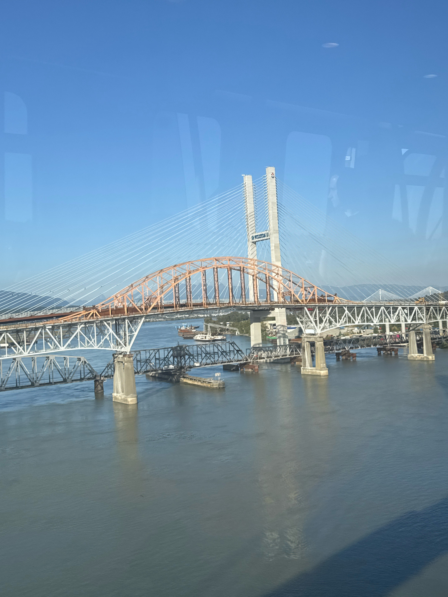 The image captures the Fraser River in New Westminster, Canada, dominated by the Port Mann Bridge and the Pattullo Bridge. The Port Mann Bridge, a cable-stayed bridge, is prominently displayed with its distinctive orange arch and tall concrete pylons. The Pattullo Bridge, a truss bridge, is situated below and to the left of the Port Mann Bridge, characterized by its intricate metal structure. The river itself appears calm, reflecting the clear blue sky. Several barges and other vessels are visible on the water, suggesting ongoing maritime activity. The surrounding landscape features a distant tree line and mountains under a bright, sunny sky. The photograph appears to have been taken from an elevated position, possibly from a helicopter or another bridge.