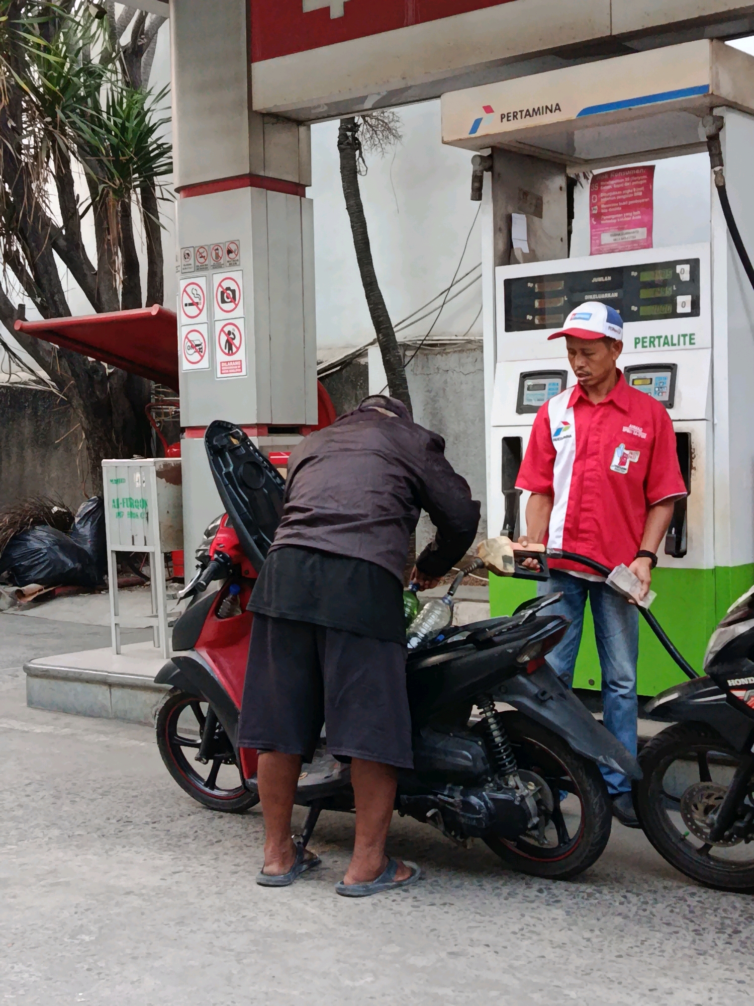 A gas station attendant wearing a red and white uniform with a white and blue cap is standing at a Pertamina gas pump in Bekasi, Indonesia. He is holding a gas nozzle in his right hand, and cash in his left. A customer, wearing a dark jacket and shorts, is standing by a red scooter, holding a plastic bottle and a green object, possibly another bottle, near the scooter's fuel tank. The gas pump displays "PERTALITE" and digital readouts of fuel quantity and price. Several no-smoking and no-photography signs are visible on the pillar of the gas station. In the background, there are trees, trash bags, and other vehicles.