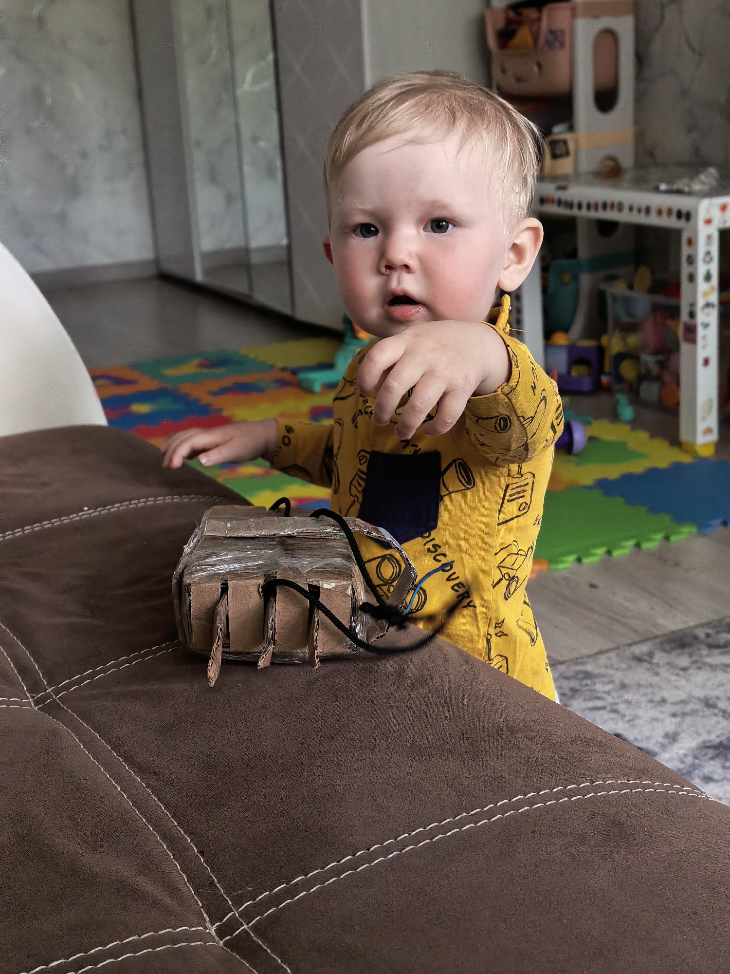 A fair-skinned, light-haired toddler stands near a brown upholstered sofa with white stitching. The child is wearing a yellow long-sleeved shirt with a dark blue patch pocket. On the sofa is a rectangular object constructed from cardboard, wrapped in clear tape, and adorned with black cords. In the background, there's a play area with colorful interlocking foam mats and a white toy shelf. The scene appears to be indoors, possibly a residential living room. The location is identified as the city of Sterlitamak, Russia.
