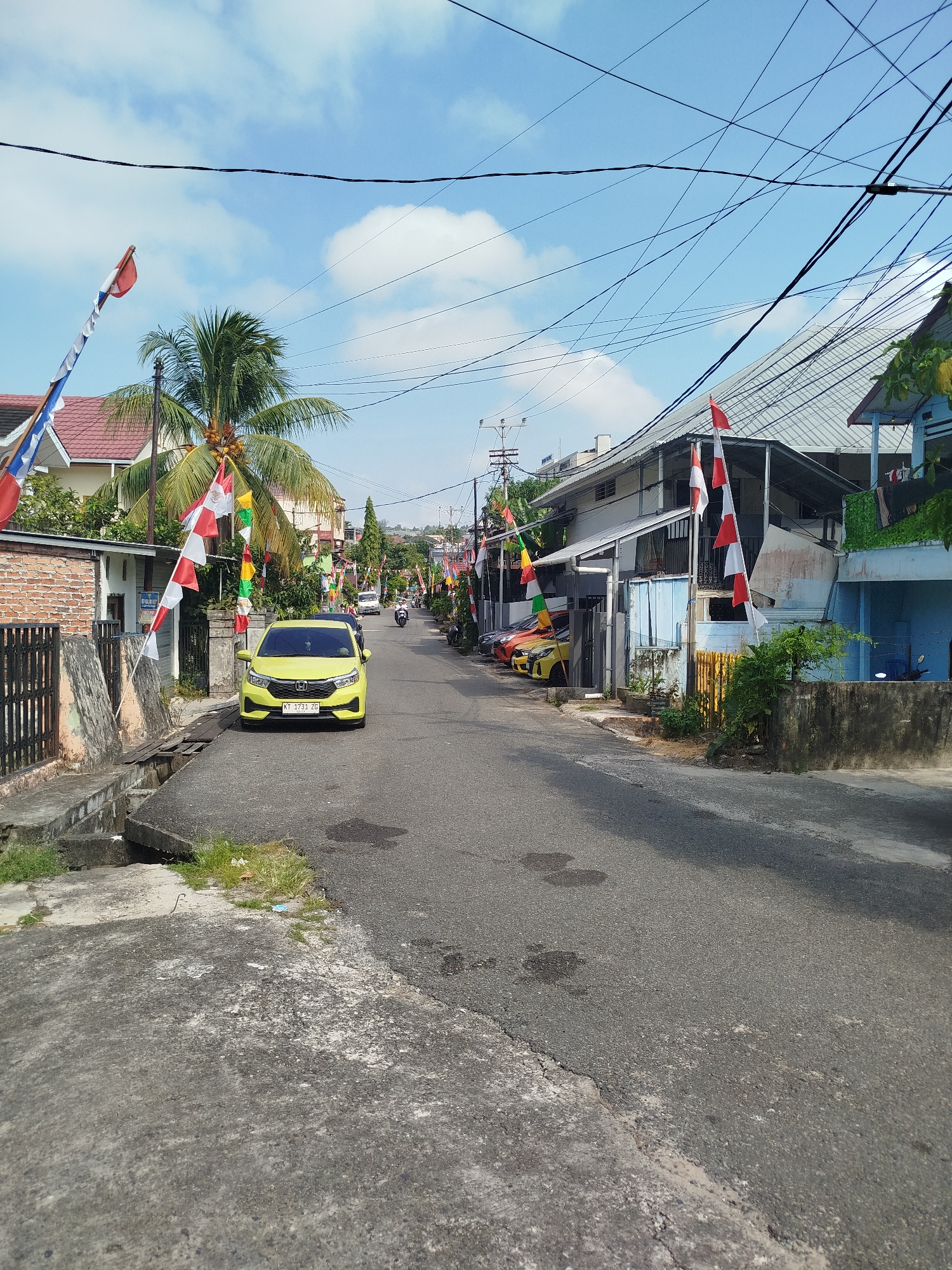 The image depicts a street scene in Balikpapan, Indonesia, on a sunny day with a partly cloudy sky. The street is paved with asphalt and has a concrete gutter on the left side. Several residential buildings line the street, some with tiled roofs and others with corrugated metal roofs. Decorative flags, predominantly red and white, are hung along the street and on poles, suggesting a festive or patriotic occasion.
A yellow car is parked on the right side of the street, with its license plate visible as "KT 1731 ZG". Several other cars are parked further down the street. A motorcycle is also visible. A palm tree with coconuts stands near the left side of the street. The environment appears to be a residential neighborhood with overhead electrical wires crisscrossing the sky. The overall impression is that of a typical Indonesian street with elements of local culture and daily life.